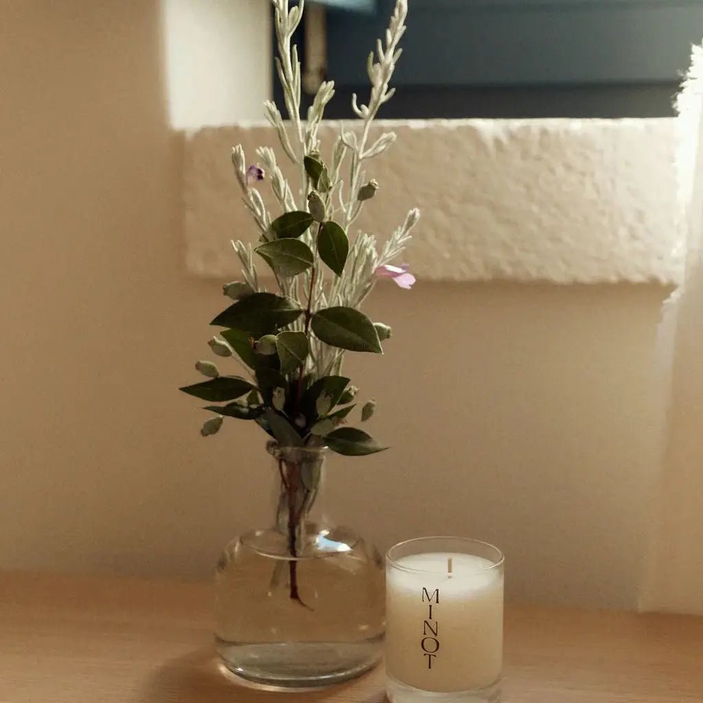 A small glass vase with green leaves and delicate flowers sits beside a Minot Luxury Non-toxic Soy Candle (3oz) on a light wooden surface in front of a window.