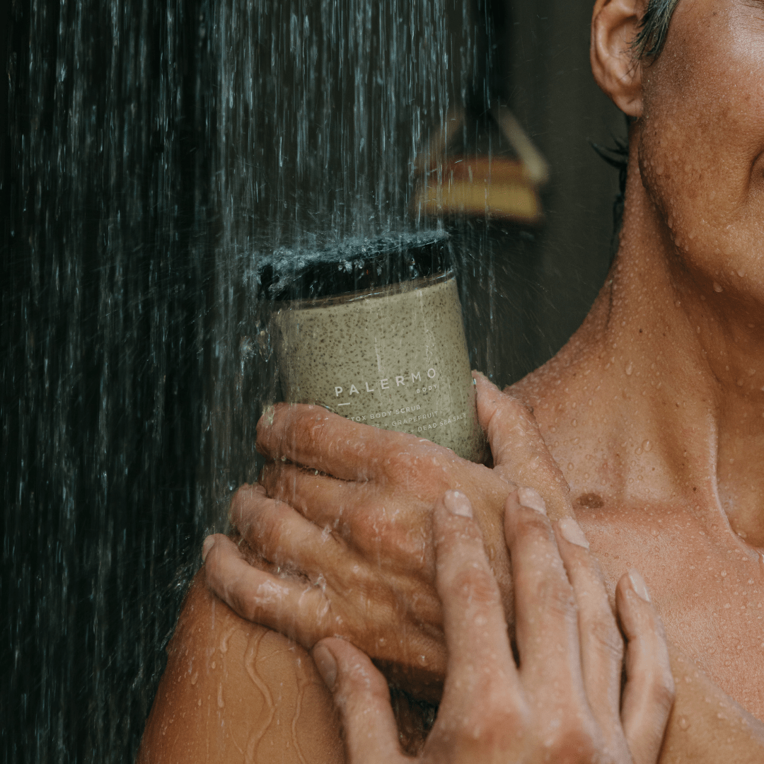 A person holds a Palermo Body Detox Body Scrub container to their chest while standing under a shower, water flowing over their shoulder and hand.