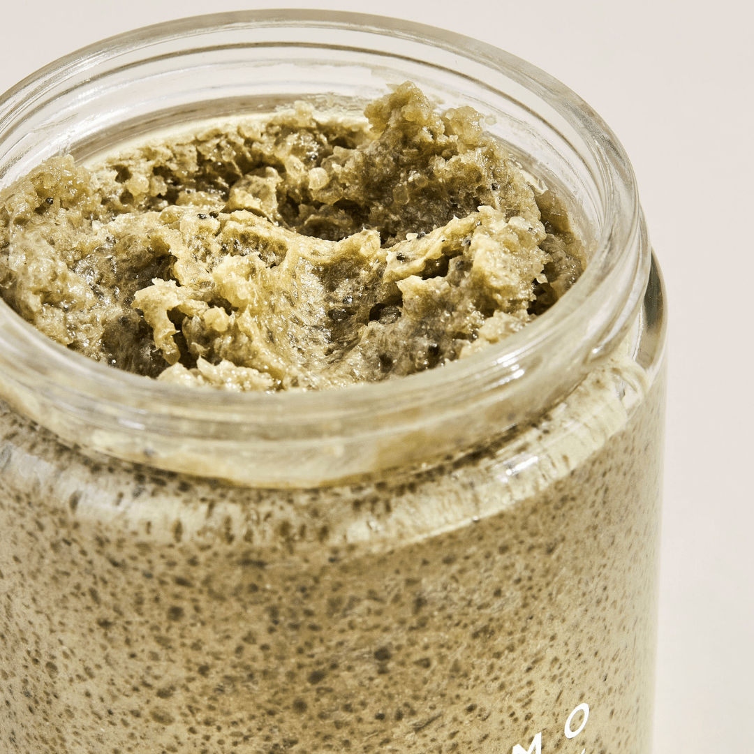 Palermo Body's Detox Body Scrub is shown in an open glass jar, revealing its thick, textured blend of sea clay and Dead Sea salt with a greenish-brown, grainy surface.
