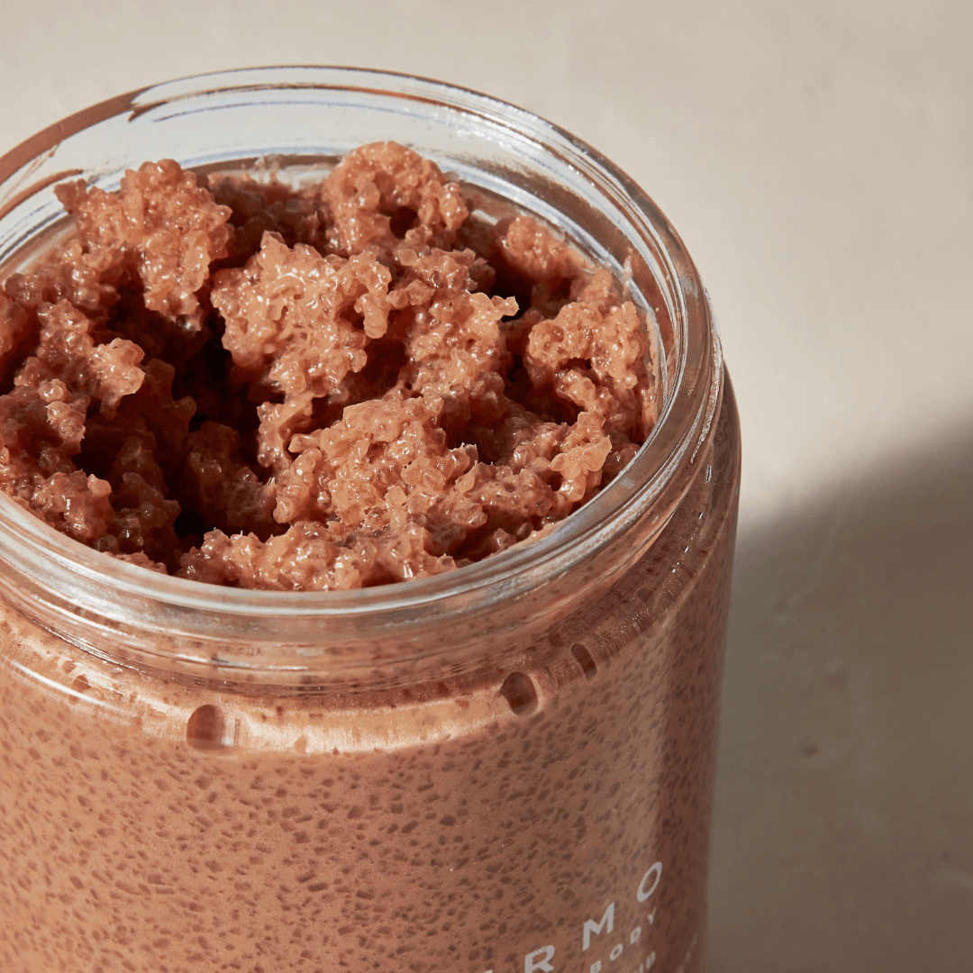 A close-up of Palermo Body’s Revitalizing Body Scrub in an open glass jar, showcasing its brown, textured formula on a light surface, smooth and inviting like French pink clay.