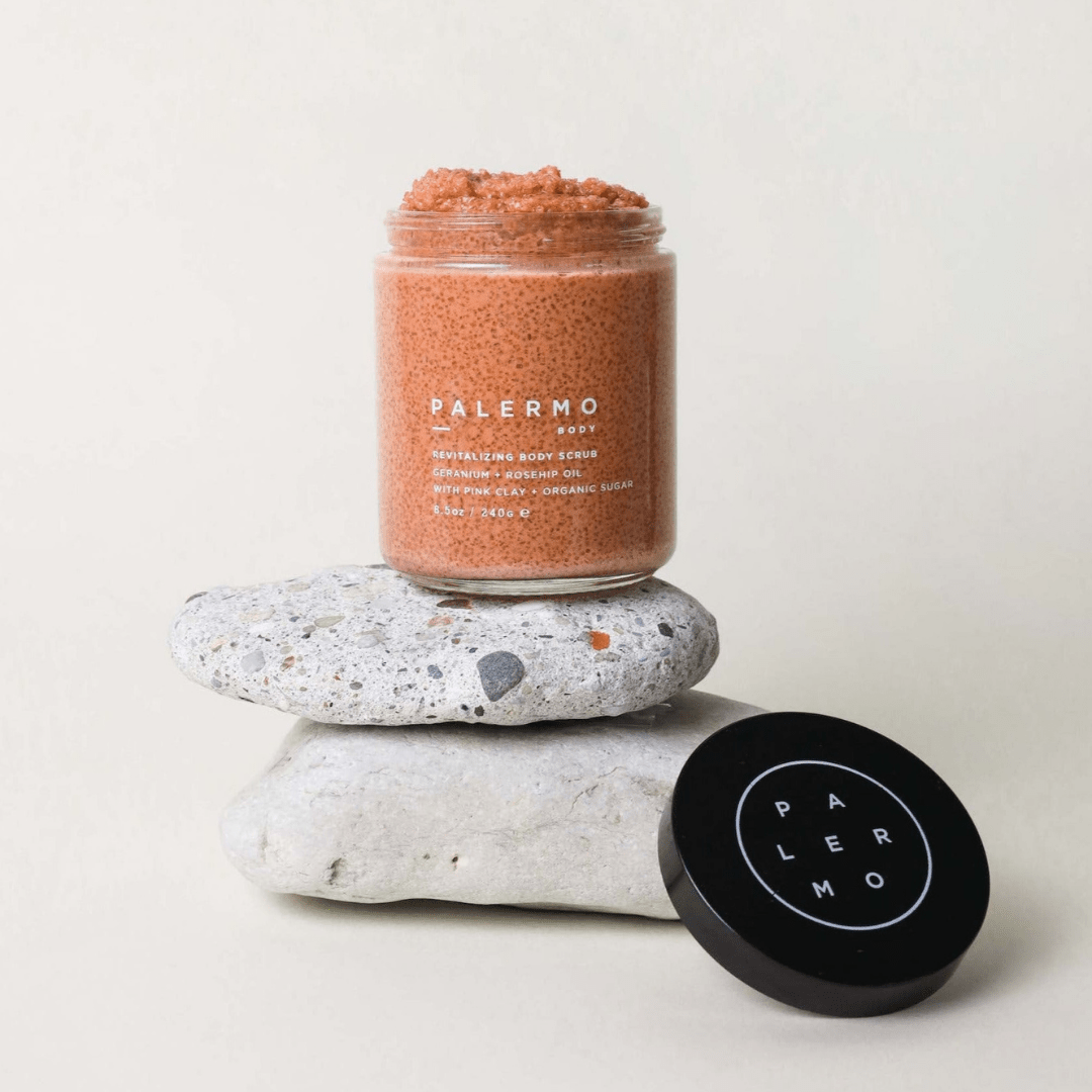 A jar of Palermo Body Revitalizing Body Scrub sits atop two stacked stones, its black "PALERMO" lid nearby on a light background.