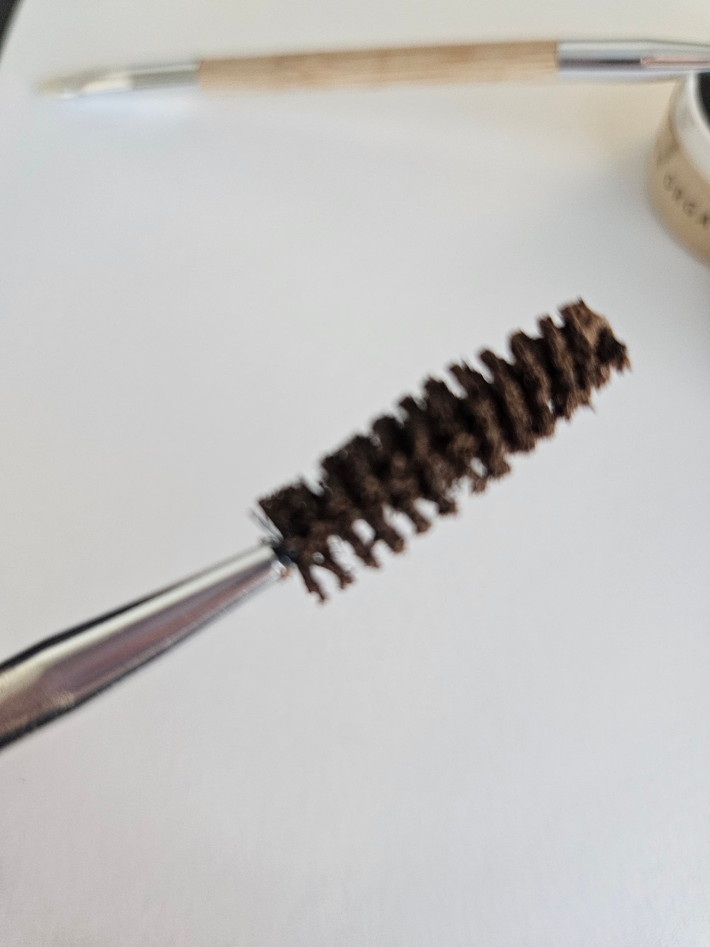 Close-up of River Organics' Eco-Friendly Double-Sided Bare Wood Brow & Mascara Brush, resembling a spoolie, held by tweezers with bristles coated in dark mascara. The background features a blurred pencil, tape spool, and white paper.