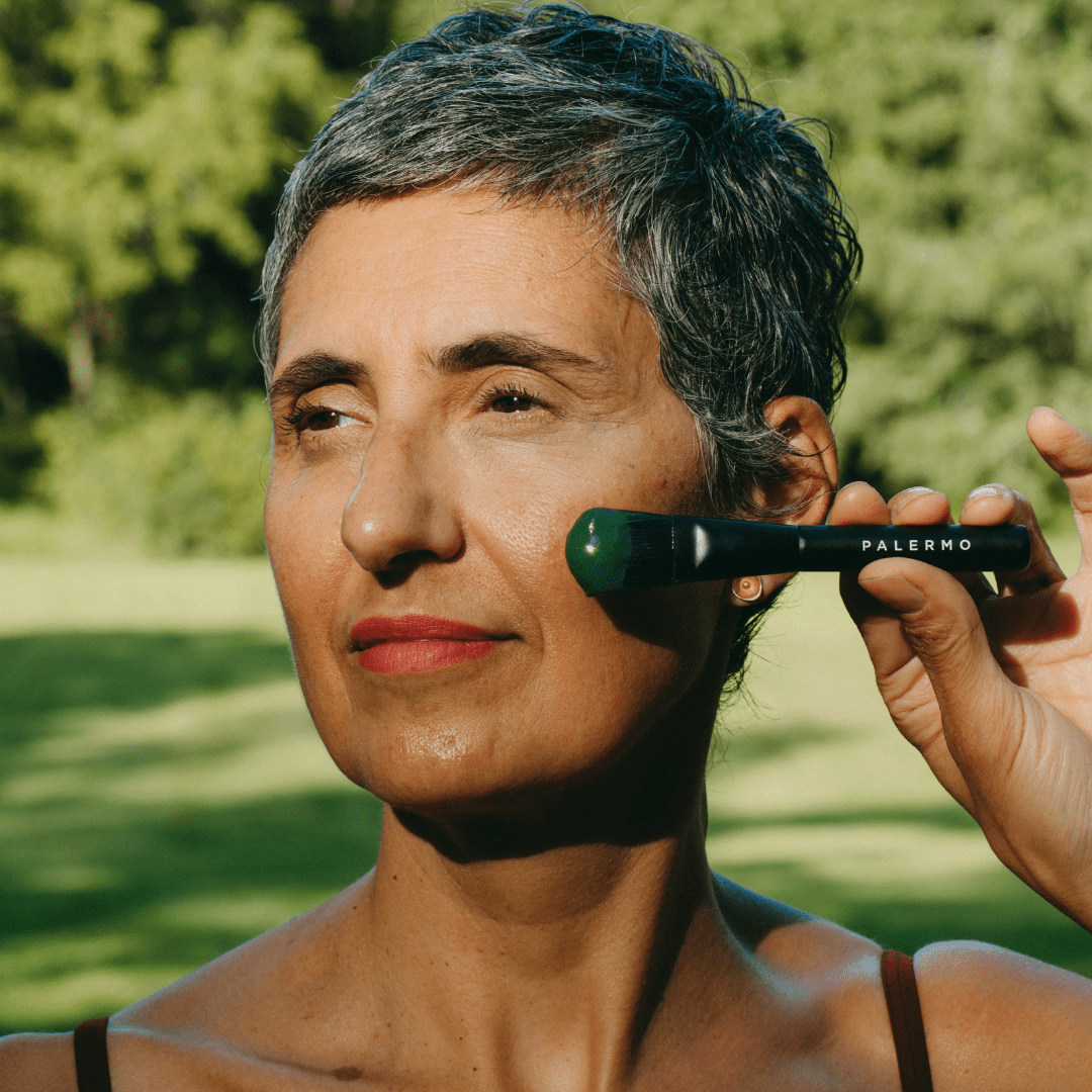 Standing outdoors in natural sunlight with greenery behind them, a person with short gray hair uses a Palermo Body facial roller on their cheek after applying the Detox Facial Mask.