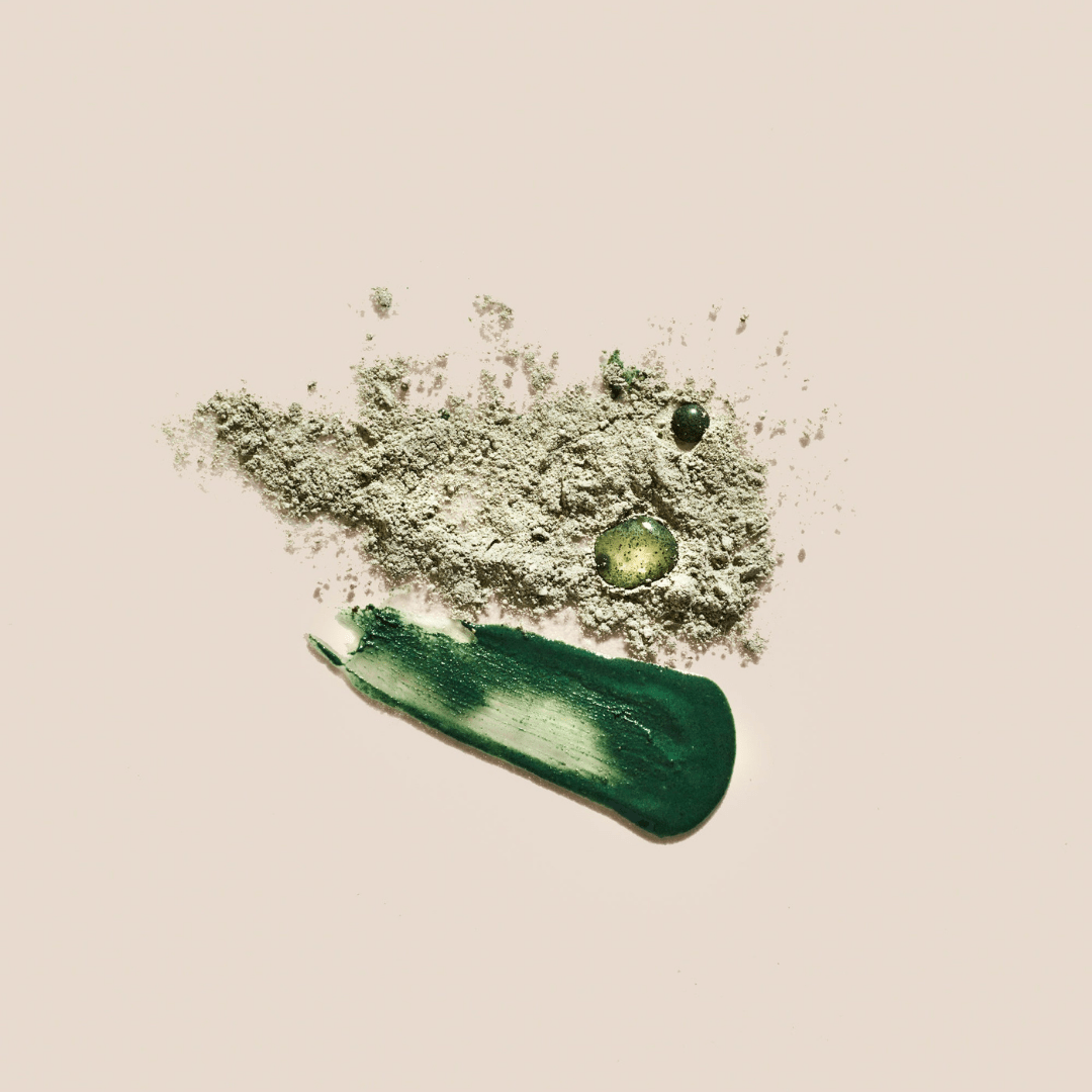 A smear of Palermo Body's Detox Facial Mask in dark green with a few drops of liquid and a light beige clarifying powder are artfully arranged on a pale background.