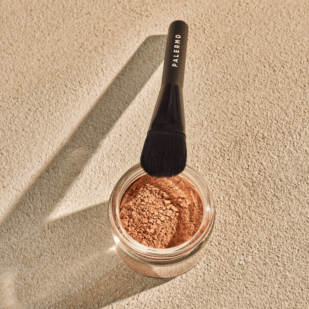 A Palermo Body makeup brush rests on a jar of Vitamin C Facial Mask—loose, light brown powder for gentle exfoliation—casting a shadow on a textured beige surface.