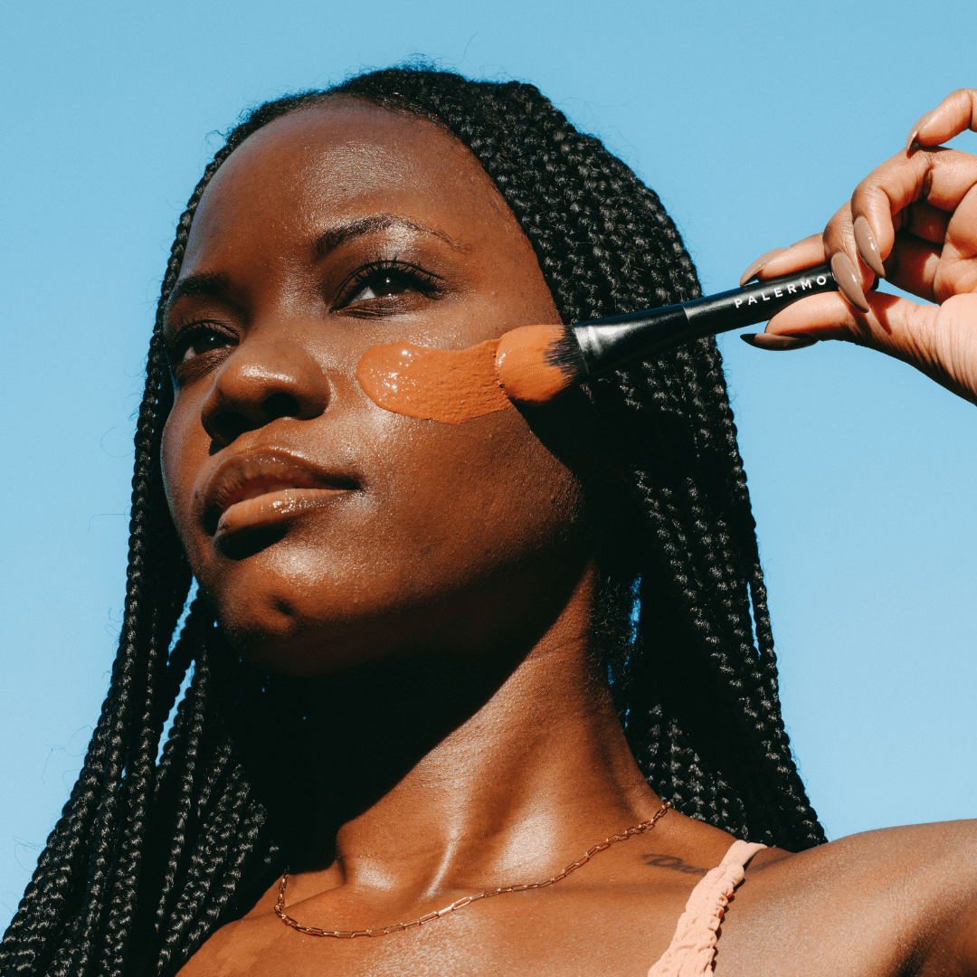 A woman with long braids applies Palermo Body’s Vitamin C Facial Mask, her skin glowing from gentle exfoliation. She wears a thin strap top and a delicate necklace beneath a clear blue sky.