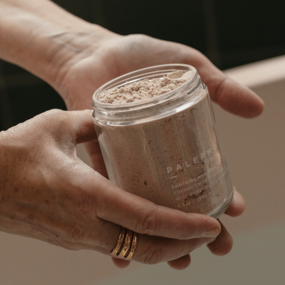 A person with a gold ring holds a glass jar of Palermo Body Soothing Milk Bath, containing light brown powder. The black background emphasizes the hands and Palermo Body’s soothing skin treatment.