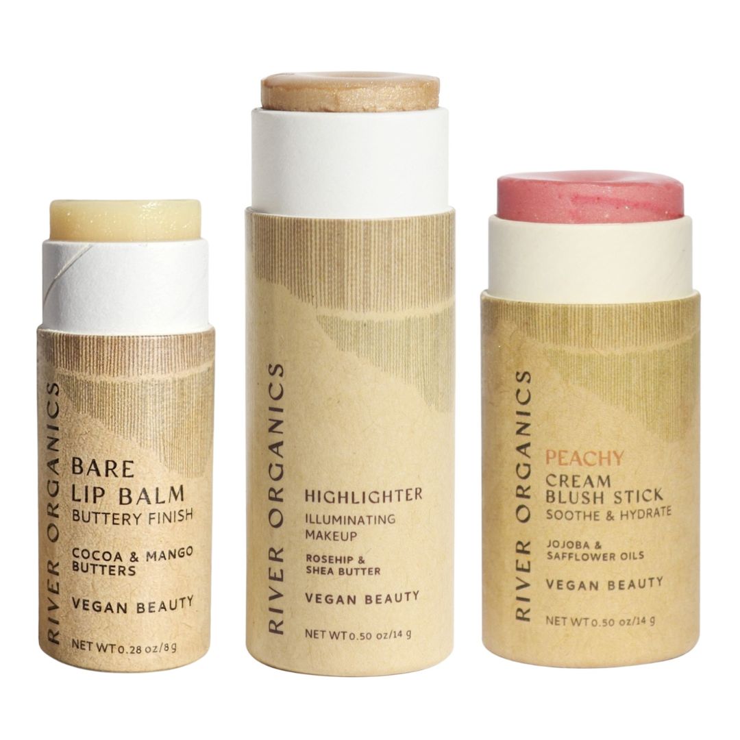 River Organics’ Lip + Highlighter + Blush Set features three cylindrical vegan beauty products in beige, plastic-free packaging, labeled with product names and ingredients—perfect for a clean beauty routine.