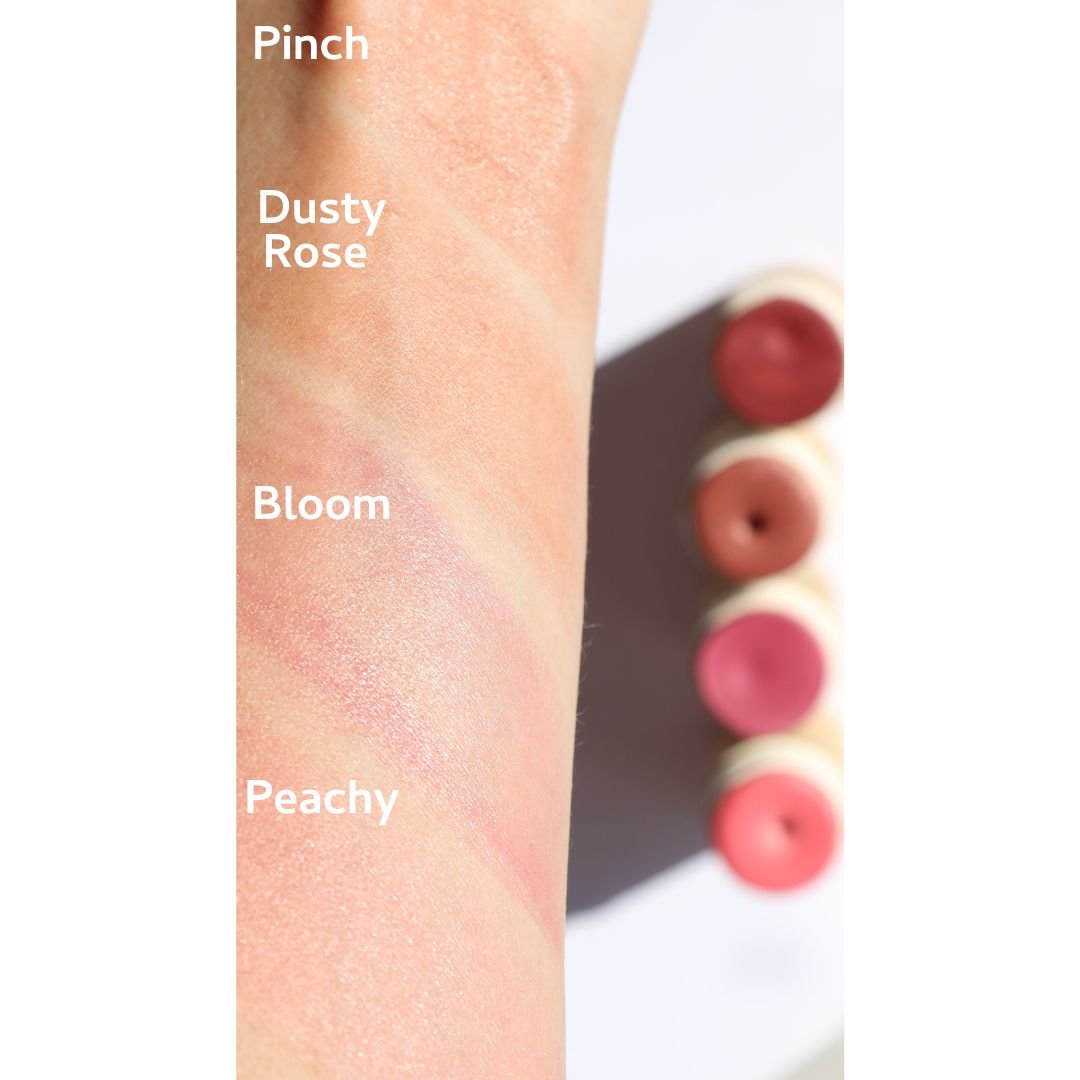 A forearm displays four swatches of River Organics' Natural Cream Blush Stick—Pinch, Dusty Rose, Bloom, and Peachy—beside matching open blush sticks in plastic-free packaging, all set against a white background.