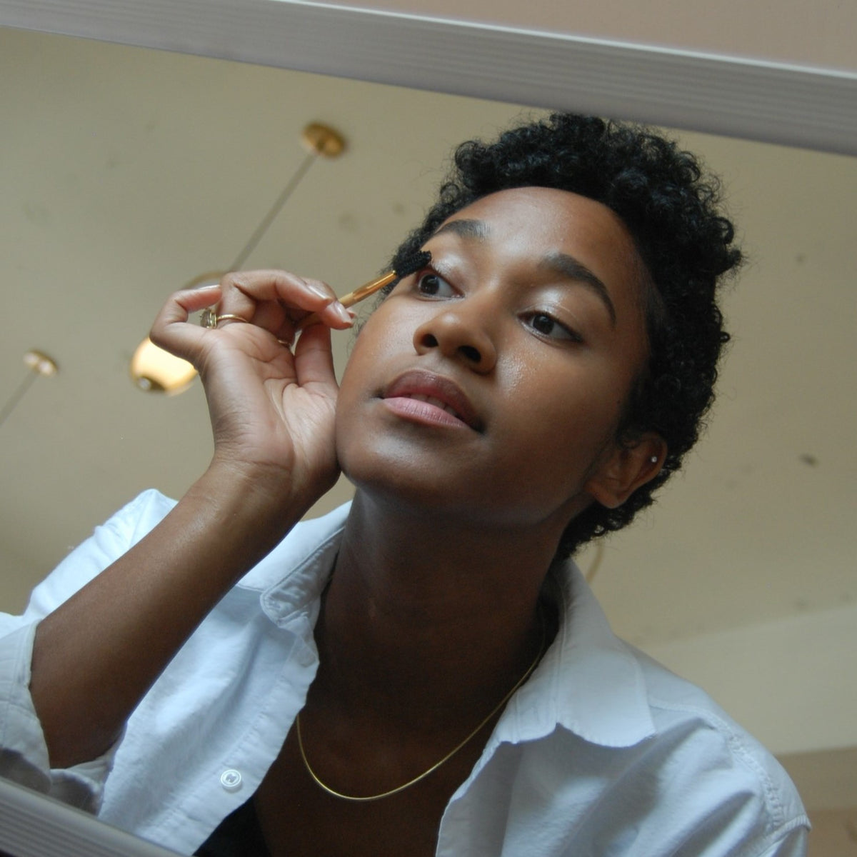 A woman with short curly hair and a white shirt applies eyeshadow while looking into a mirror. Warm lights, a neutral ceiling, and her River Organics Black Vegan Zero-Waste Mascara are reflected in the background.