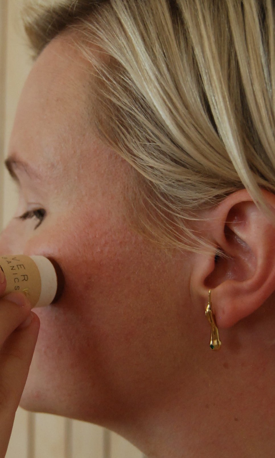 A person with short blonde hair applies River Organics Creamy Vegan Bronzing Stick to their cheek for a sun-kissed glow, wearing a gold twig-shaped earring.