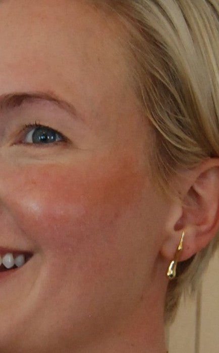 A smiling person with short blonde hair, blue eye, and light skin shows off a sun-kissed glow using River Organics' Creamy Vegan Bronzing Stick. Wearing a gold drop earring, only the left side of their face is visible.
