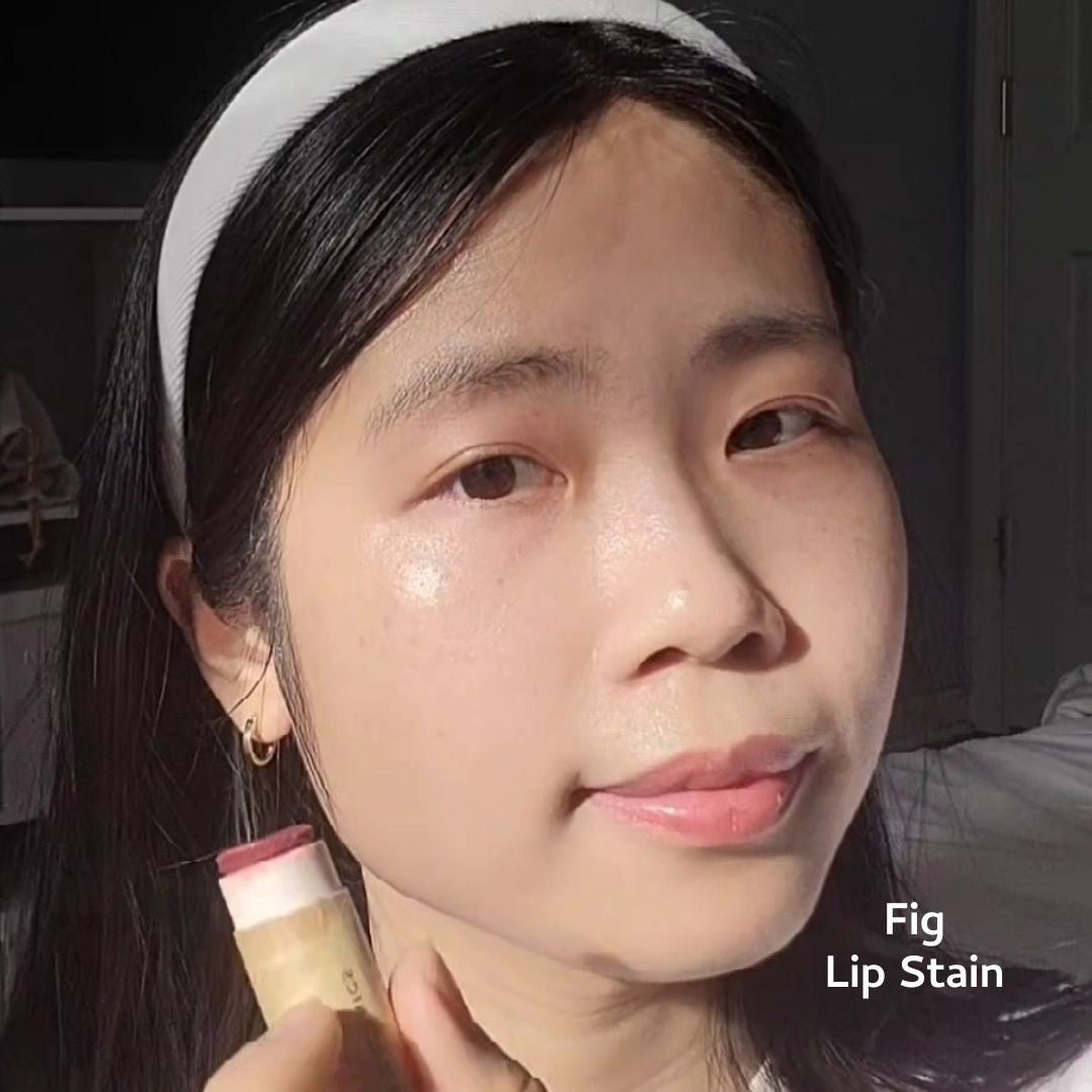 A woman with straight black hair and a white headband applies River Organics Vegan Lip Stains to her lips. "Fig Lip Stain" appears in the lower right corner, and she is in a bright, sunlit room.