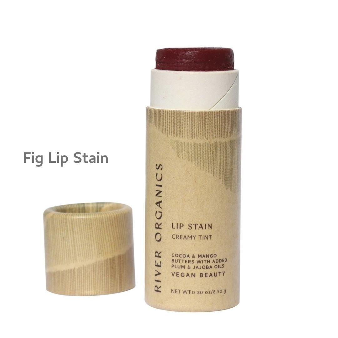 An open tube of River Organics Vegan Lip Stains sits with its cardboard cap beside it. The label notes hydrating cocoa and mango butters, plum and jojoba oils, emphasizing vegan, plastic-free beauty in a deep red natural tint.