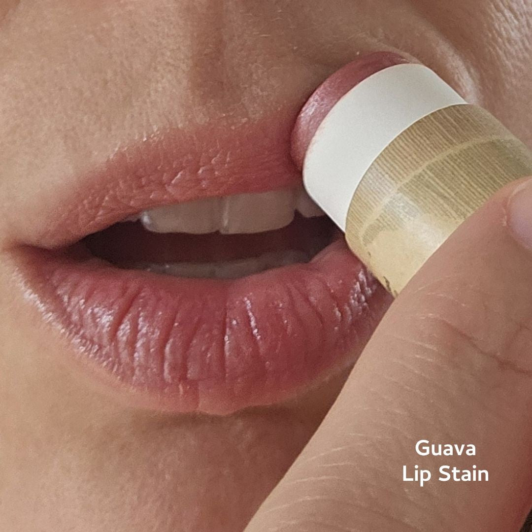 A close-up of someone applying River Organics' Vegan Lip Stains—a natural pinkish tint—to their lips. Their mouth and teeth are visible, and the beige-and-white tube highlights the brand's chic, eco-friendly design.