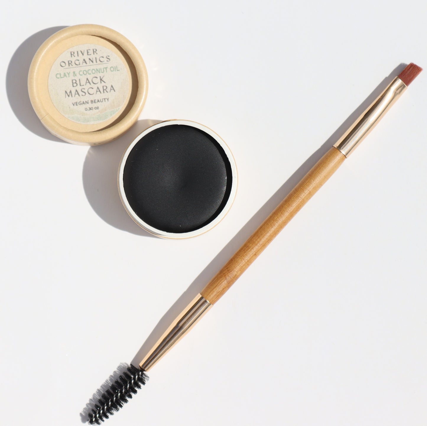 An open River Organics clay and coconut oil black mascara sits ready for application beside an Eco-Friendly Double-Sided Bare Wood Brow & Mascara Brush, featuring a spoolie and angled end, all against a crisp white background.