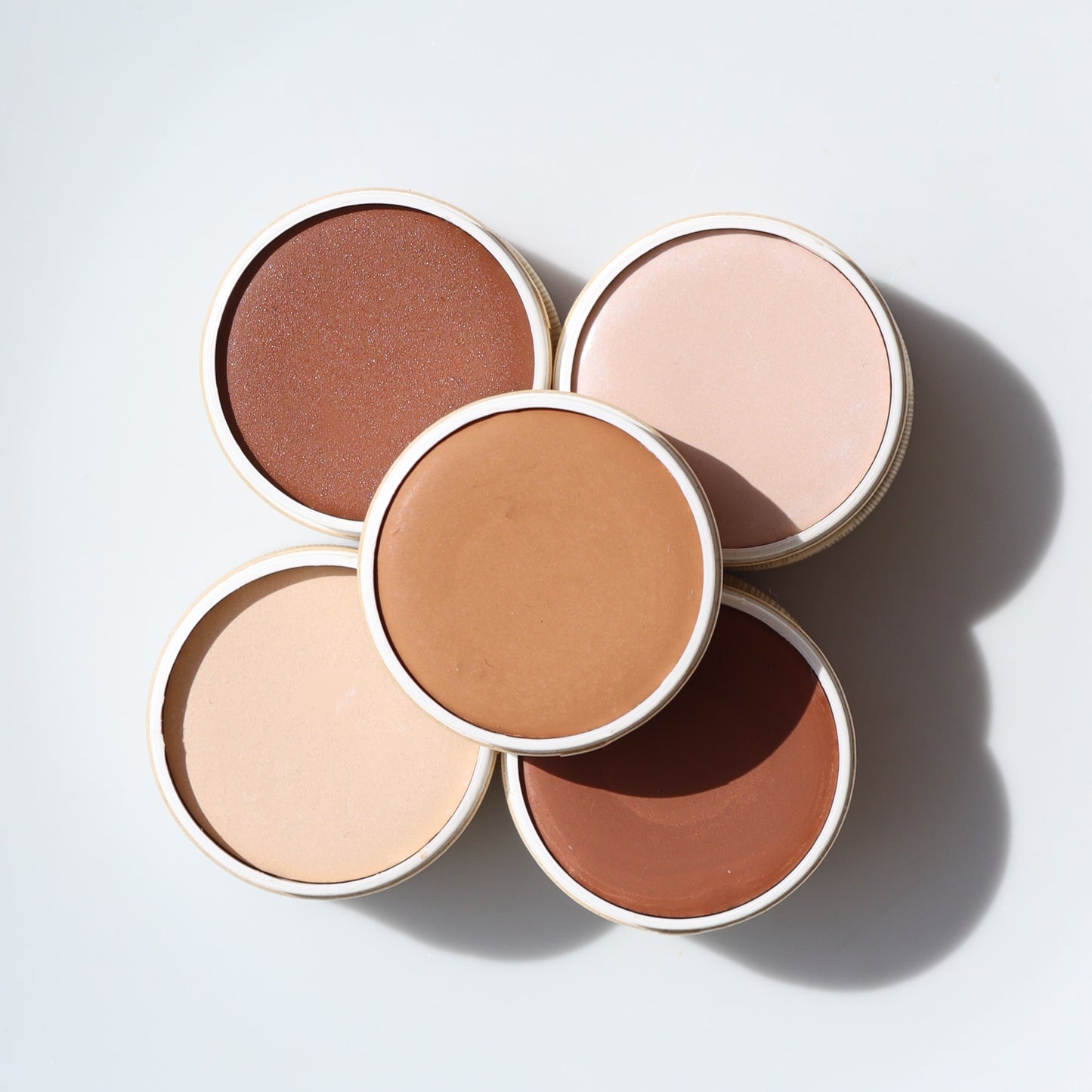 Five round containers of River Organics Zero-Waste Vegan Concealer in various skin tone shades are arranged in a flower-like pattern on a white surface.
