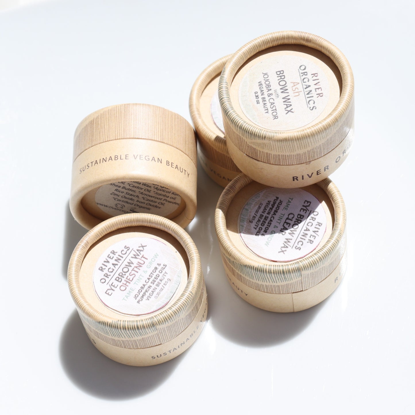 Five small, round wooden containers of River Organics Brow Wax - Tame, Tint & Grow rest on a white surface. Each is labeled "Sustainable Vegan Beauty" with product info on top as bright sunlight casts shadows.