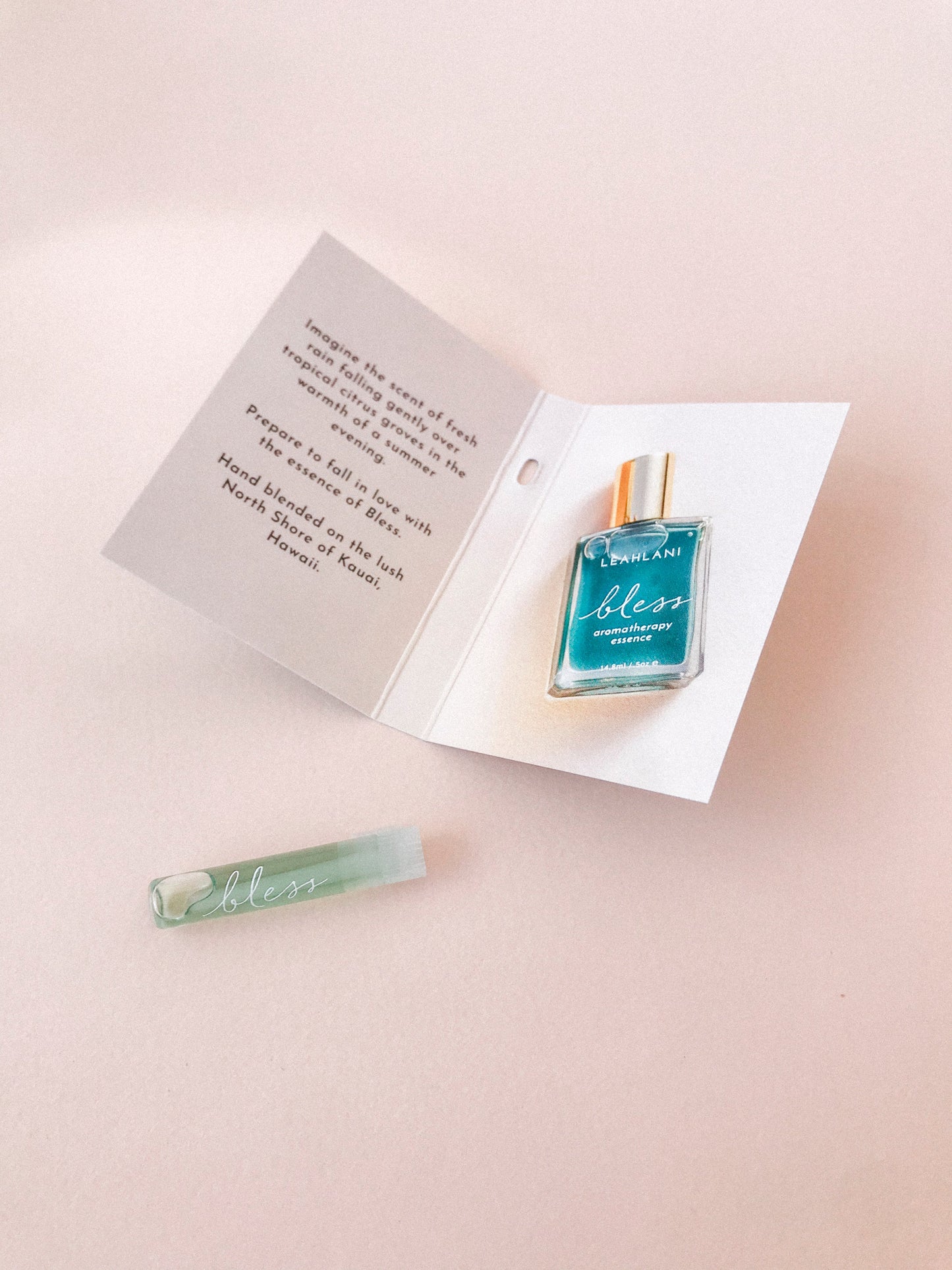 A small turquoise bottle of LEAHLANI's Bless Aromatherapy Essence and a sample vial are displayed on a pale surface beside an open card featuring printed details about this tropical, citrus fragrance.