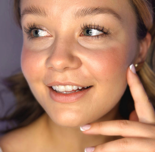A woman with light makeup smiles softly, touching her cheek with a manicured hand as she applies the River Organics Natural Cream Blush Stick. Her complexion appears clear, her lips are glossy, and her eyebrows are styled. The softly blurred background draws attention to her radiant face and gentle expression.