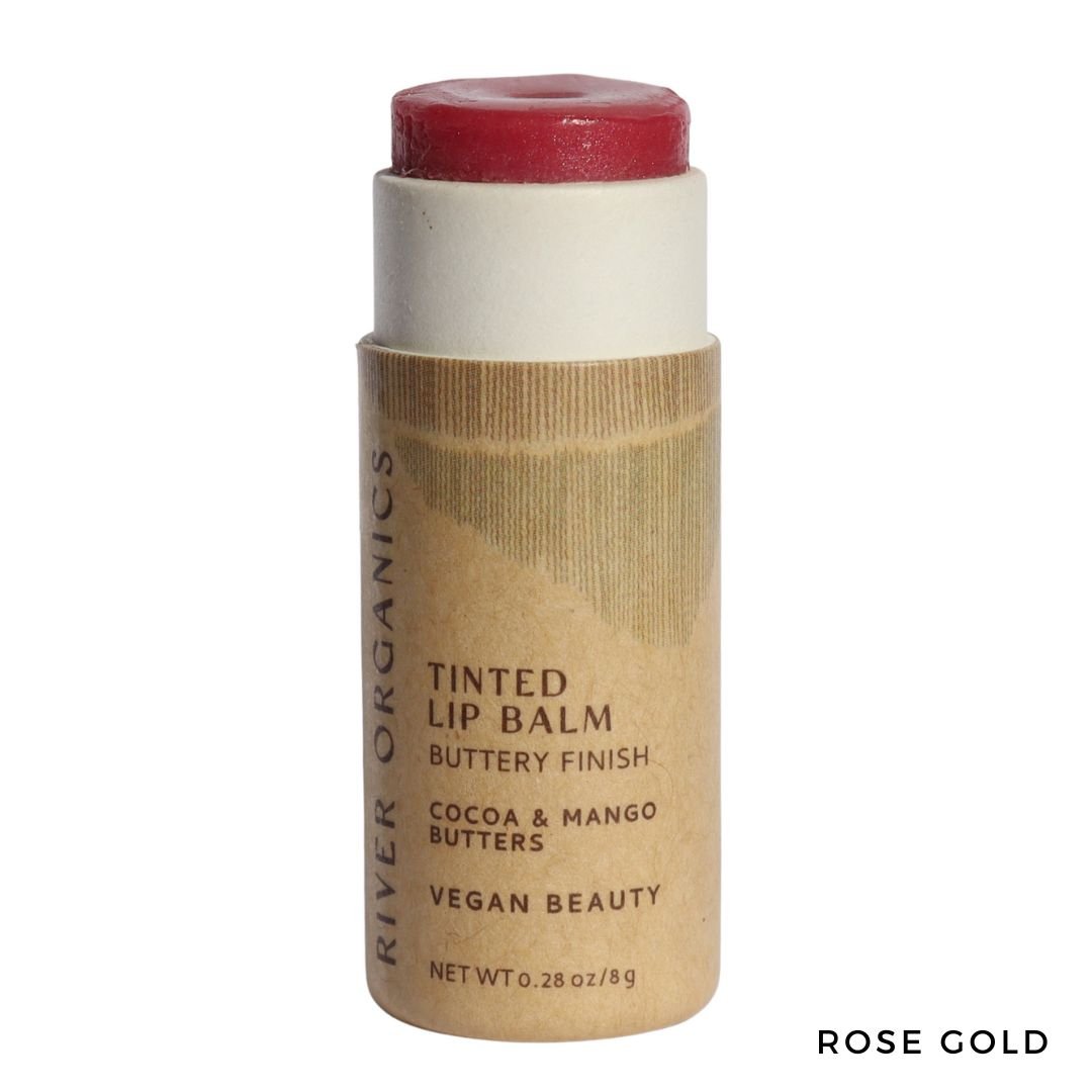 A tube of River Organics Tinted Vegan Lip Balm in rose gold with cardboard packaging. The label features plant-based cocoa and mango butters for deep moisture and a buttery vegan finish. The balm is partially raised above the tube.