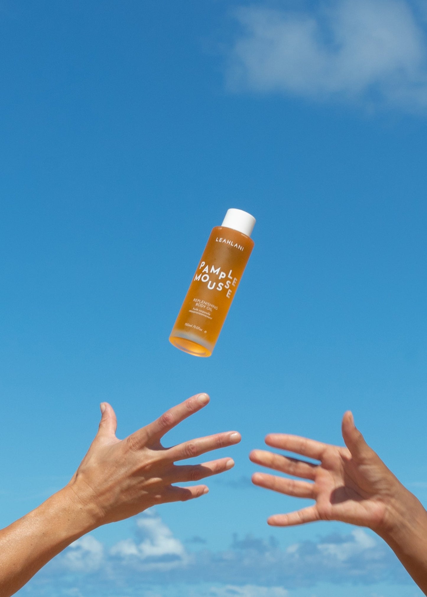 A bottle of LEAHLANI Pamplemousse Replenishing Body Oil floats against a bright blue sky as two hands reach up toward it from below.
