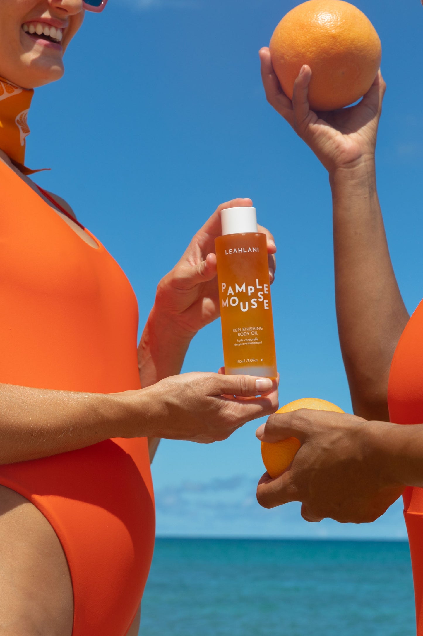 Two people in orange swimsuits hold grapefruits and a bottle of LEAHLANI Pamplemousse Replenishing Body Oil, featuring tropical oils, against a bright blue sky and ocean for a vibrant, summery look.