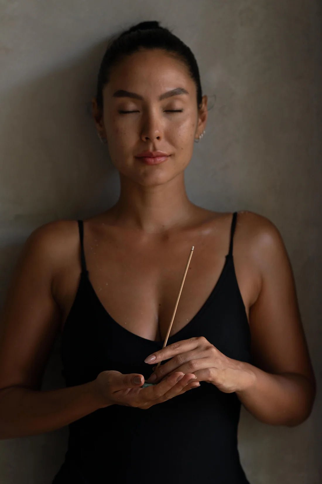 A woman with closed eyes stands against a gray wall, holding a lit Nusa Holistick Essential Oil Incense - Lotus, Champaka in both hands, wearing a black tank top and appearing calm and meditative.