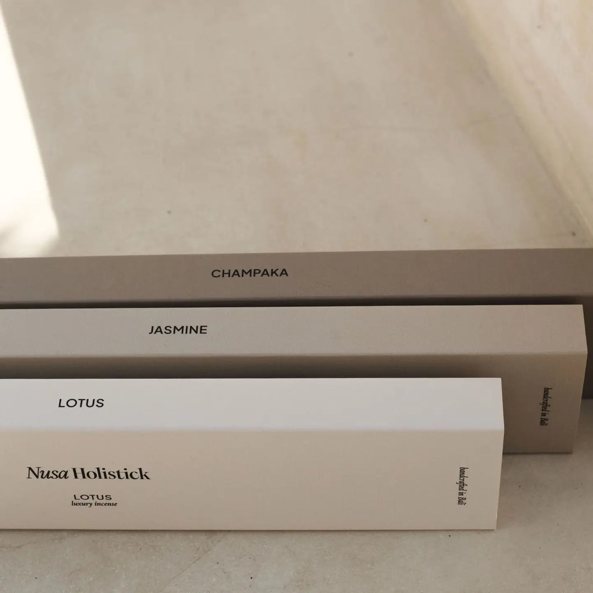 Three rectangular incense boxes labeled “CHAMPAKA,” “JASMINE,” and “LOTUS” from Nusa Holistick are stacked on a smooth beige surface, with “Nusa Holistick incense” and “Lotus luxury incense” displayed on the front box. Blended with Balinese essential oils and inspired by the gentle essence of the lotus flower, these Essential Oil Incense sticks in Lotus and Champaka create a serene ambiance in your space.