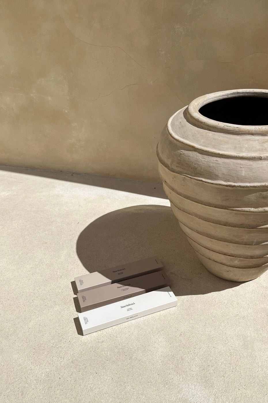A large, textured beige pottery vase casts a shadow on a sunlit floor. Beside it, three rectangular boxes of Nusa Holistick’s Essential Oil Incense in Lotus and Champaka are neatly arranged side by side in neutral hues.