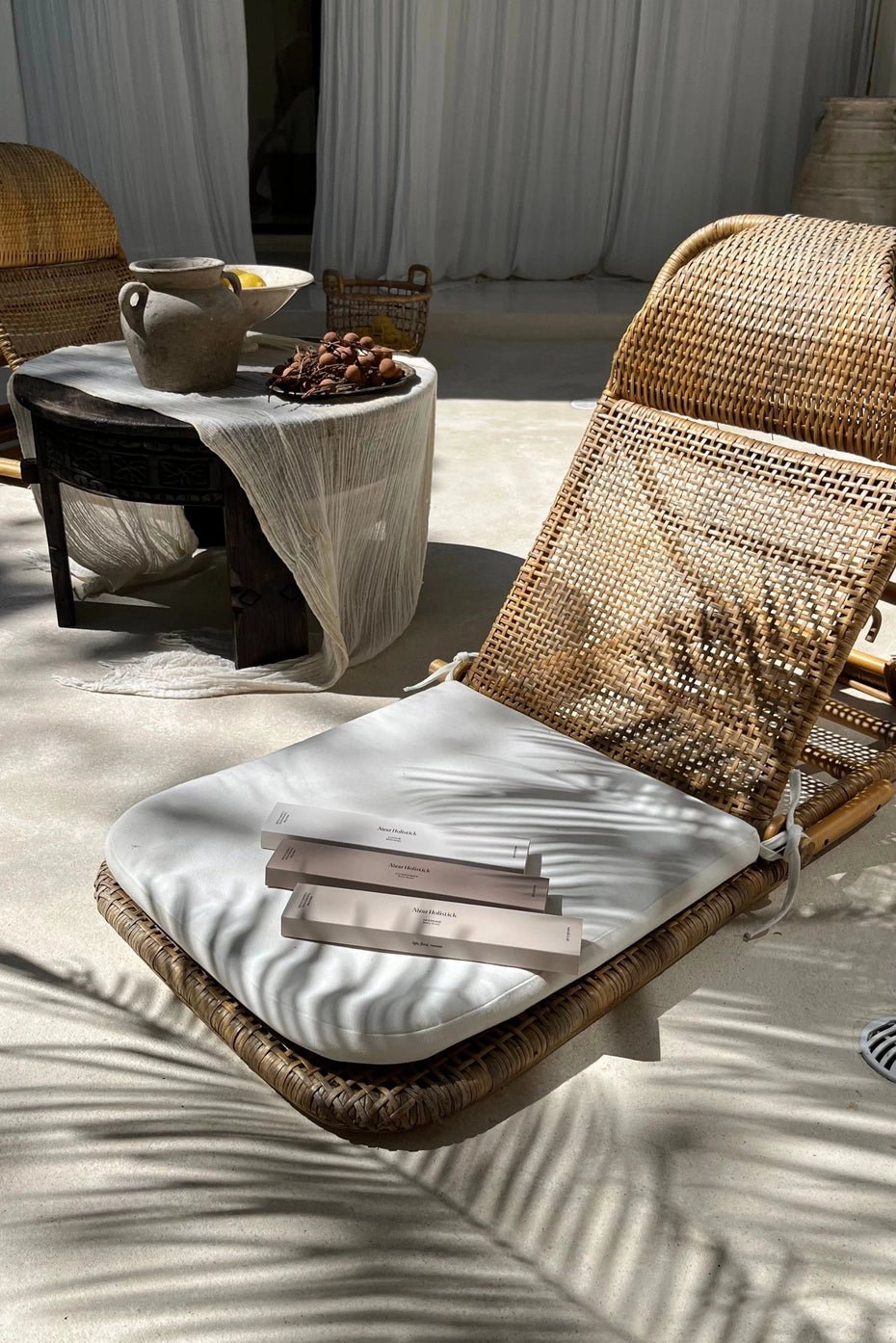 A wicker lounge chair with a white cushion holds three skincare product boxes, while a Nusa Holistick Essential Oil Incense in Lotus and Champaka rests delicately on the armrest. Nearby, a small table draped in white cloth displays a ceramic jug, bowl, and nuts. Soft sunlight casts gentle shadows across the patio, filling the scene with subtle hints of Balinese-inspired essential oils.