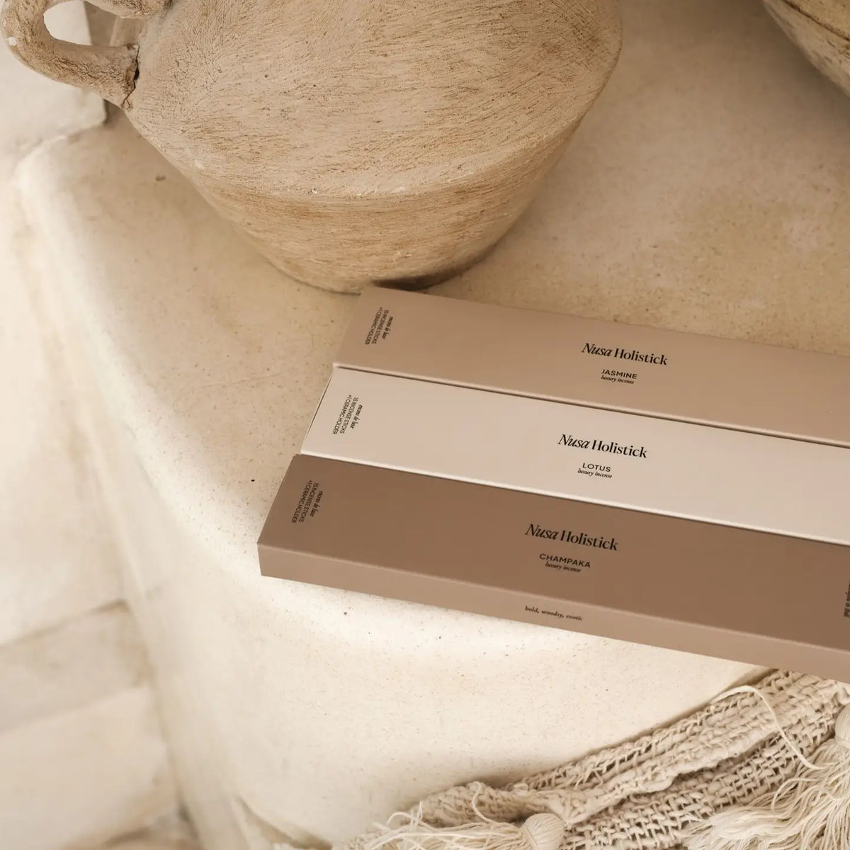 Three rectangular boxes of Nusa Holistick Essential Oil Incense—Lotus and Champaka—in soft beige tones rest on a stone or plaster surface beside a woven bag and a rustic clay pot, creating an earthy, natural aesthetic inspired by Balinese essential oils.