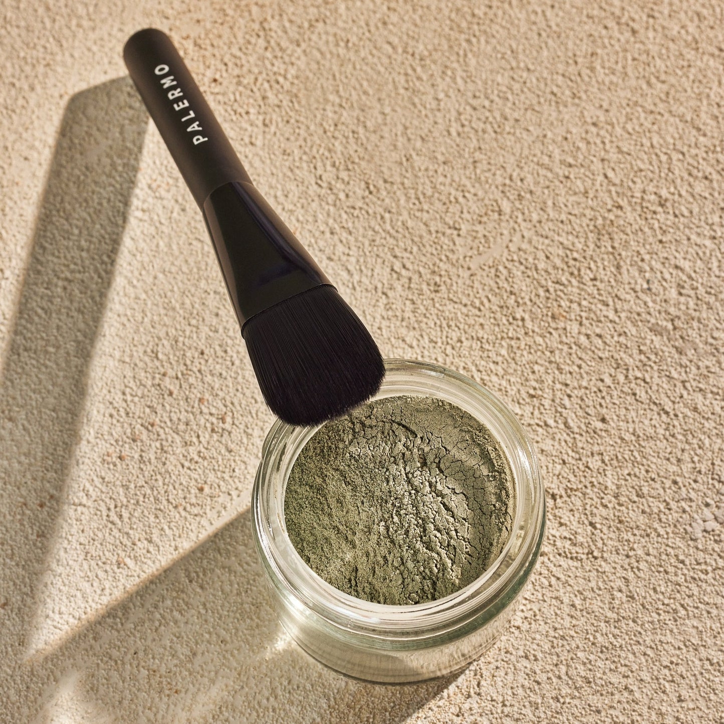 A black brush labeled "Palermo Body" rests on the rim of a glass jar filled with green Detox Facial Mask powder, set on a textured beige surface in natural light.