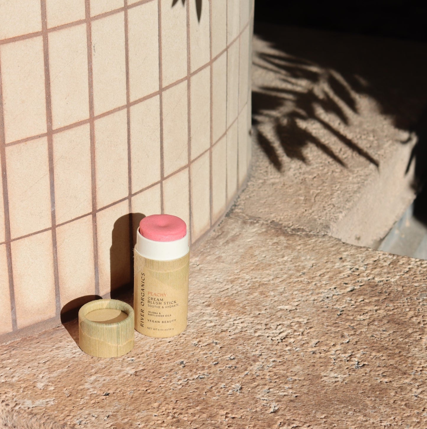 The Creamy Vegan Blush Stick - Peachy by River Organics stands upright on a textured stone surface with its cap removed. Nearby plant shadows dance on the stone, enhancing the natural ambiance of this zero-waste, partially used sustainable beauty product.