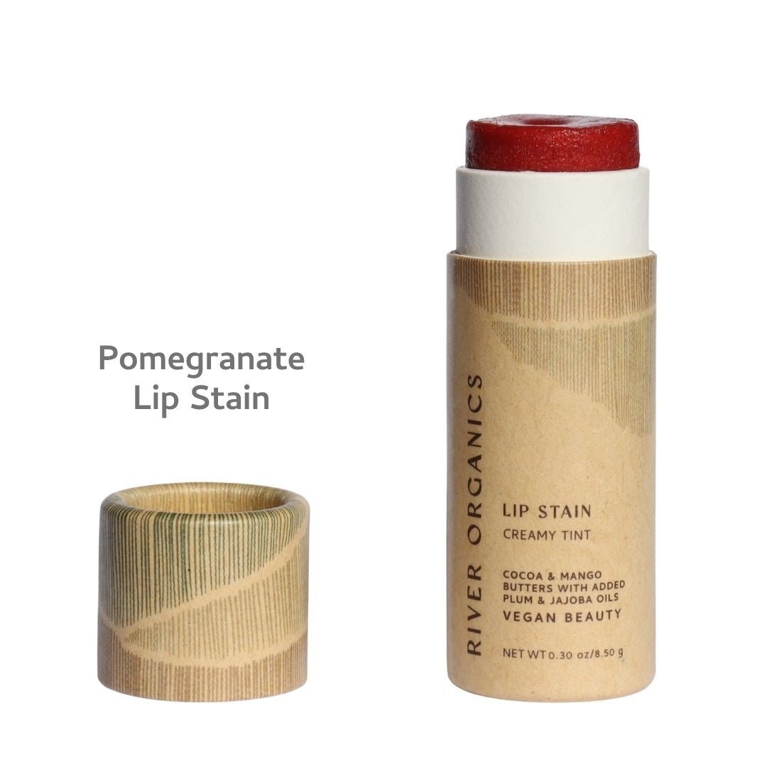 An open tube of River Organics Lip Stain: Vegan, Plastic-Free, & Organic sits with its wooden cap beside it. The packaging describes this vegan, plastic-free lip stain formulated with organic cocoa butter, mango butter, and nourishing oils. “Pomegranate Lip Stain” is printed on the label, emphasizing a long-lasting, hydrating formula designed for organic beauty enthusiasts.
