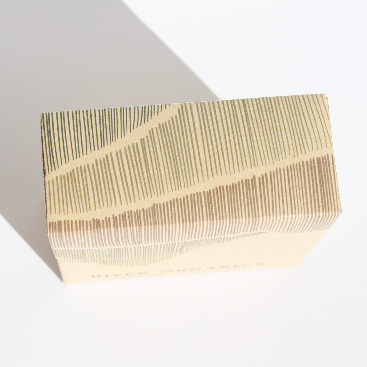 The River Organics Gift Box features a beige design with abstract brown and beige lines, casting a shadow on a white surface. It arrives fully assembled, inviting elegance and ready to contain the perfect surprise inside.