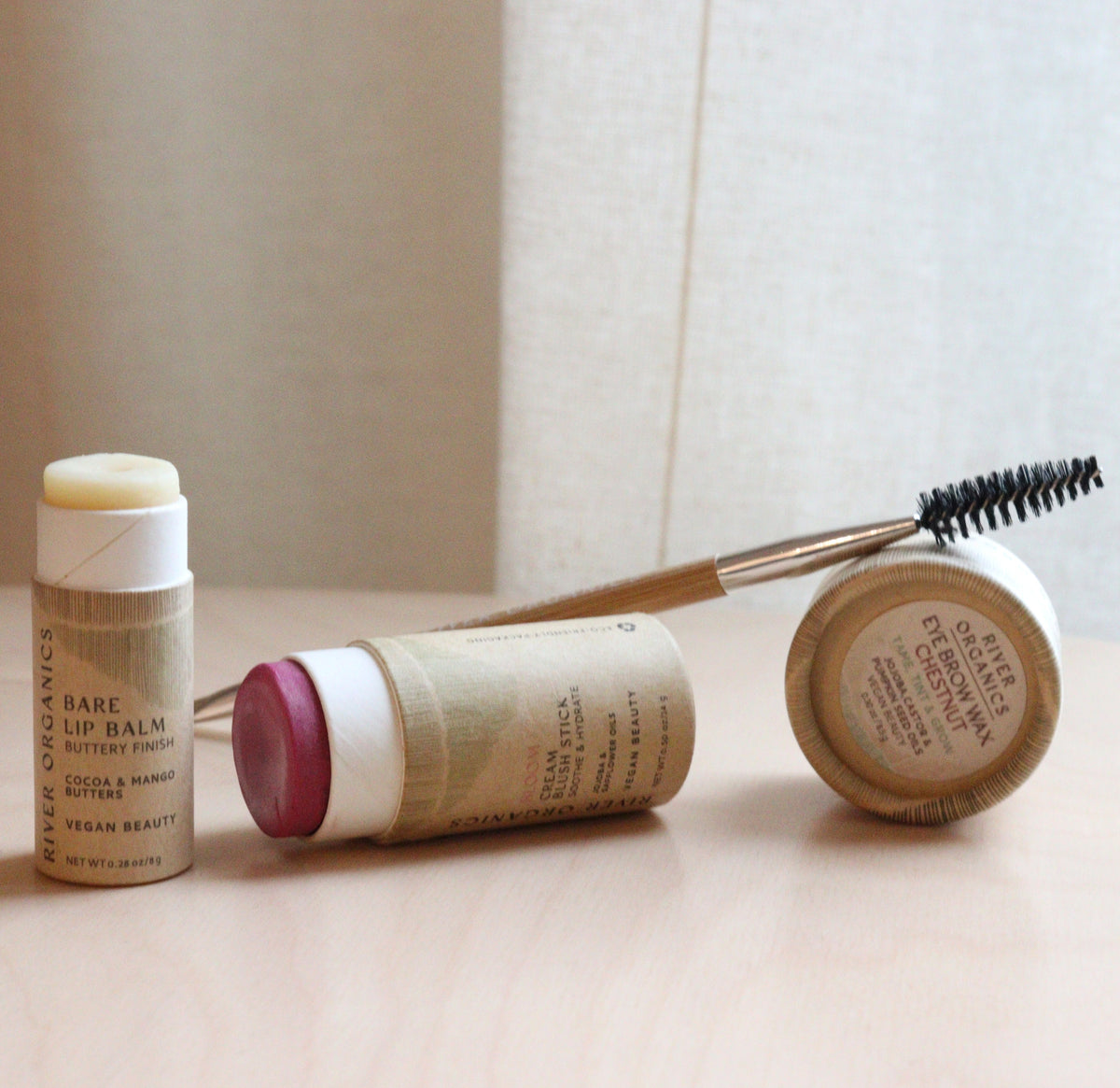 The River Organics Brow Wax + Blush + Lip Balm Bundle features three natural makeup products in eco-friendly, beige-toned packaging displayed on a wooden surface.