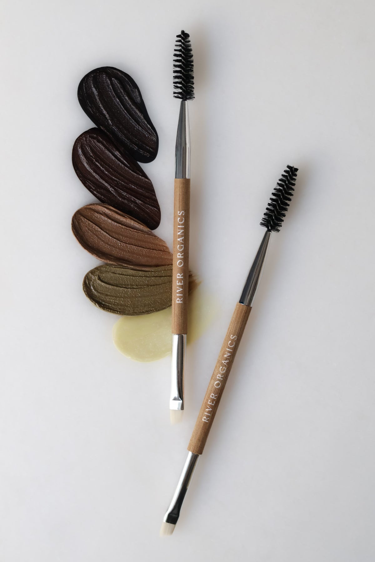 Two bamboo-handled brushes with spoolie and angled tips rest beside five brow pomade swatches in dark brown to light beige shades—the perfect companions to the River Organics Brow Wax + Blush + Lip Balm Bundle.