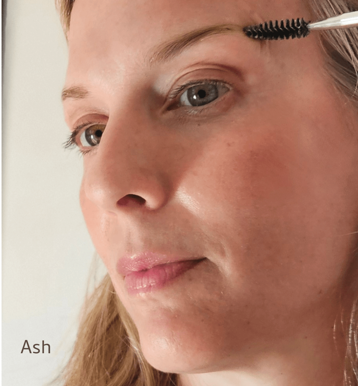 A close-up of a woman brushing her eyebrow with a spoolie, featuring River Organics Brow Wax & Growth Serum - All Colors (vegan). The word "Ash" appears in the lower left corner.