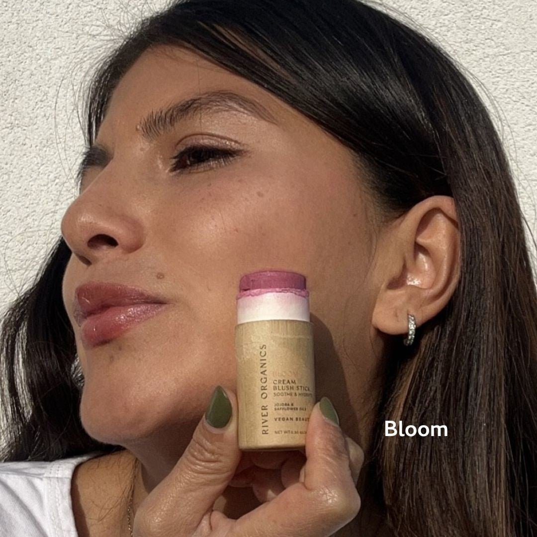 A woman with smooth skin, green-painted nails, and hoop earrings holds a River Organics Creamy Vegan Blush Stick | Set of 2 Mix & Match near her cheek in its pink shade, as “Bloom” appears in white text on the image.