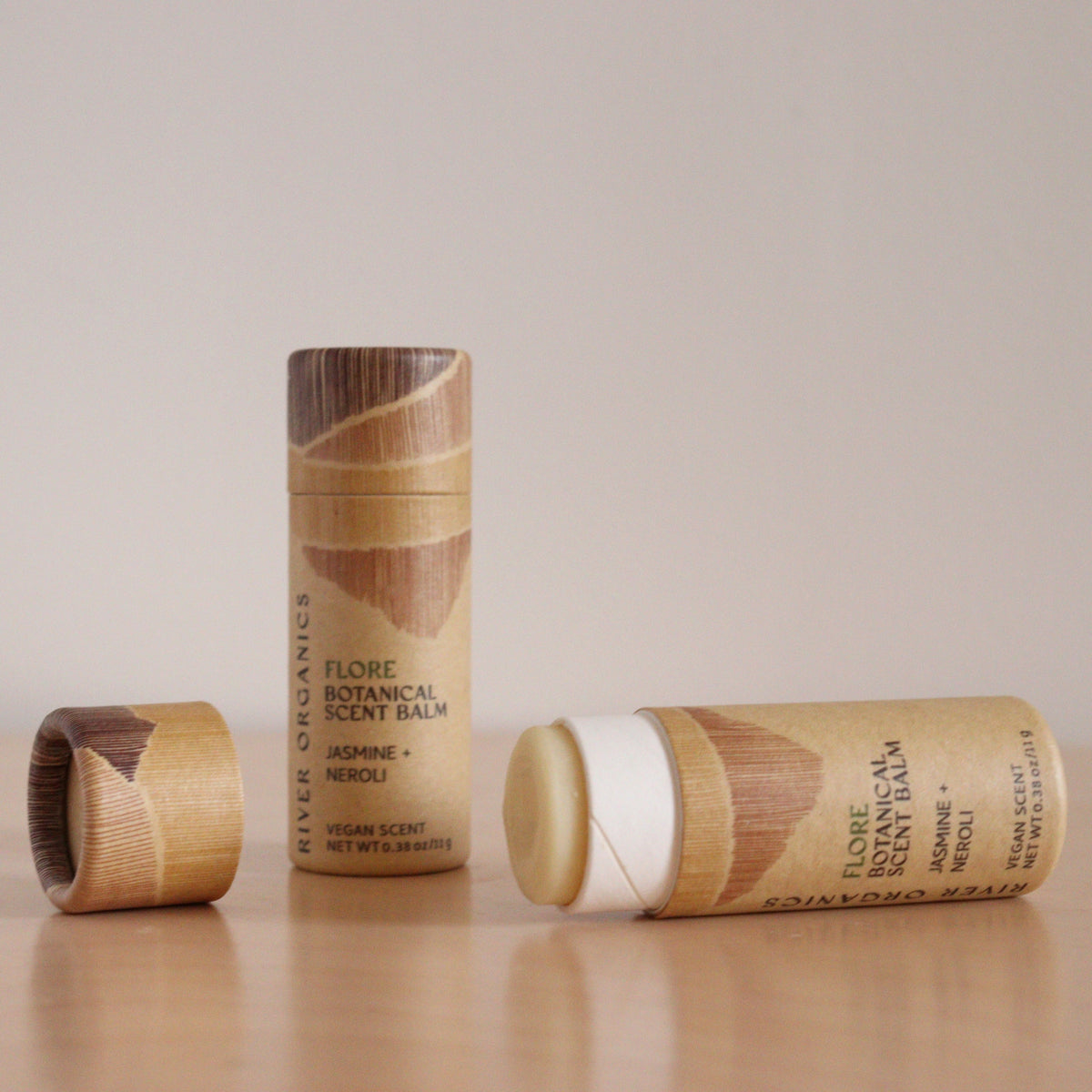 Two cylindrical tubes of River Organics Flore: Jasmine + Neroli Botanical Scent Balm are shown on a beige surface—one upright with the cap off, the other lying down with balm exposed—highlighting natural jasmine and neroli fragrance notes.