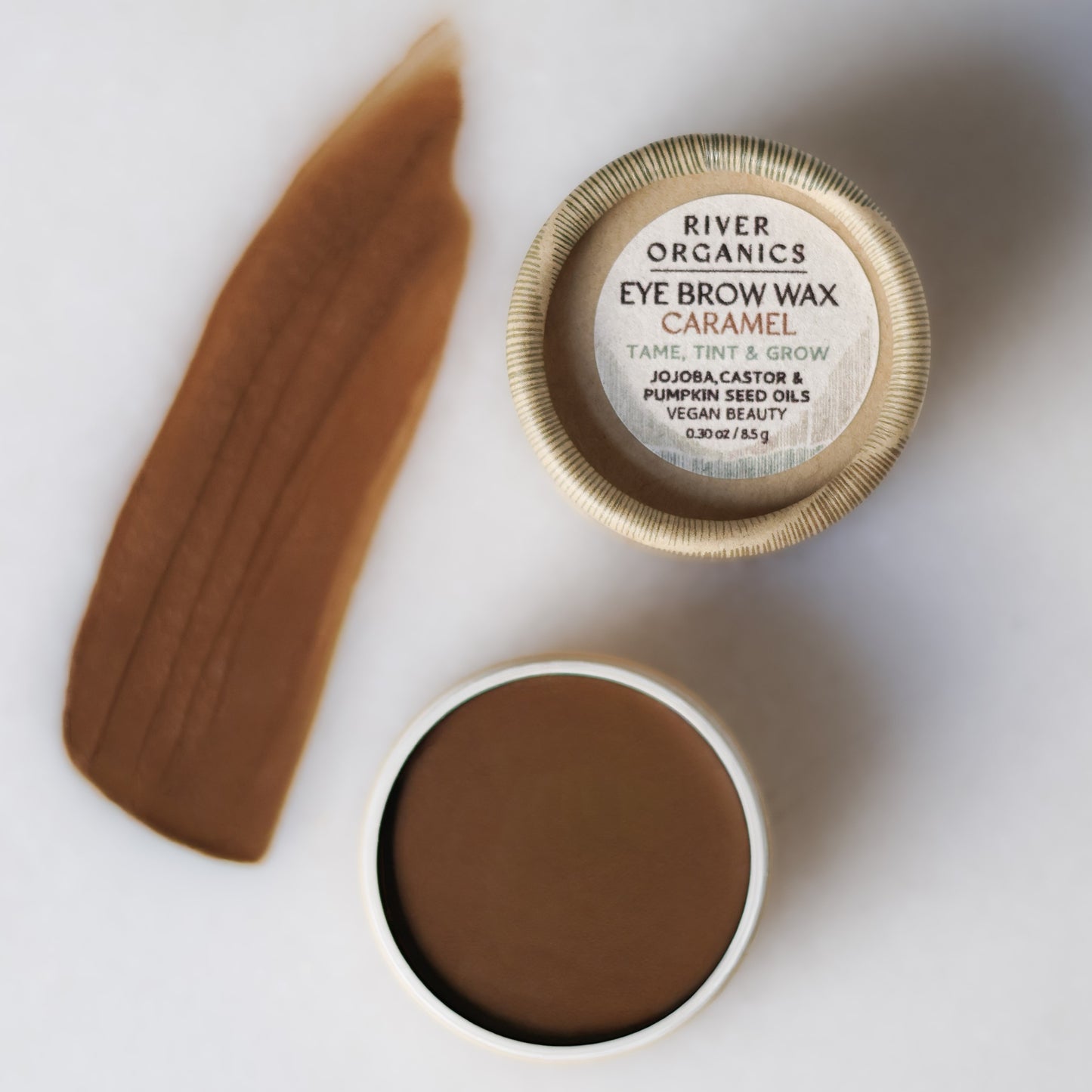 Open container of River Organics Brow Wax - Tame, Tint & Grow in Caramel, with a brown swatch on white. The label highlights jojoba, castor, and pumpkin seed oils for sustainable beauty.