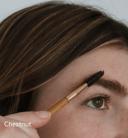 A person with brown hair grooms their brow using the River Organics Concealer & Brow Duo - Zero-Waste Vegan Beauty Set, featuring a spoolie brush and brow wax. The word "Chestnut" appears in the lower left corner.