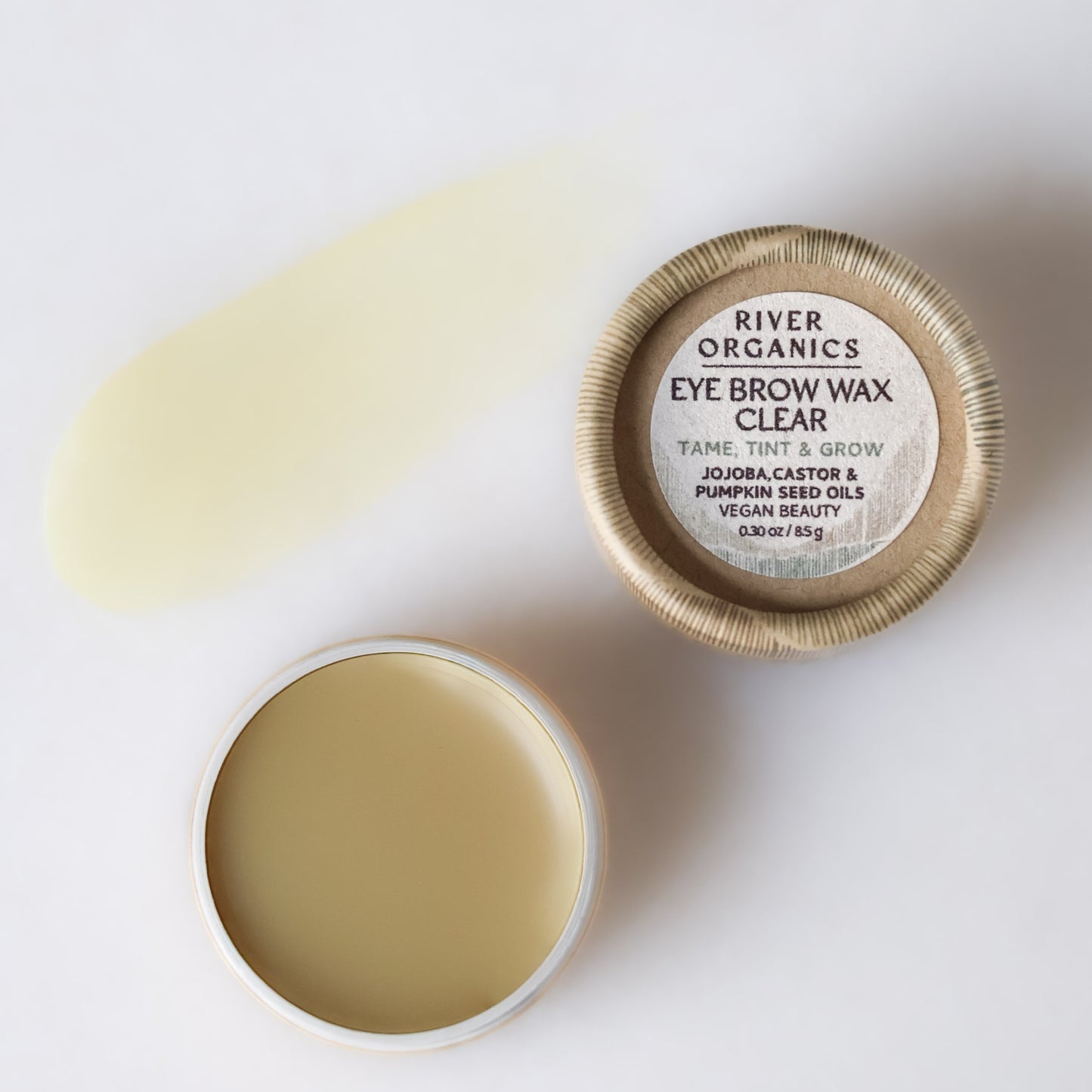An open tin of River Organics Brow Wax & Growth Serum with smoothed wax sits next to its lid. A swatch of the brow wax is spread on the white background beside the tin, showcasing its smooth texture.