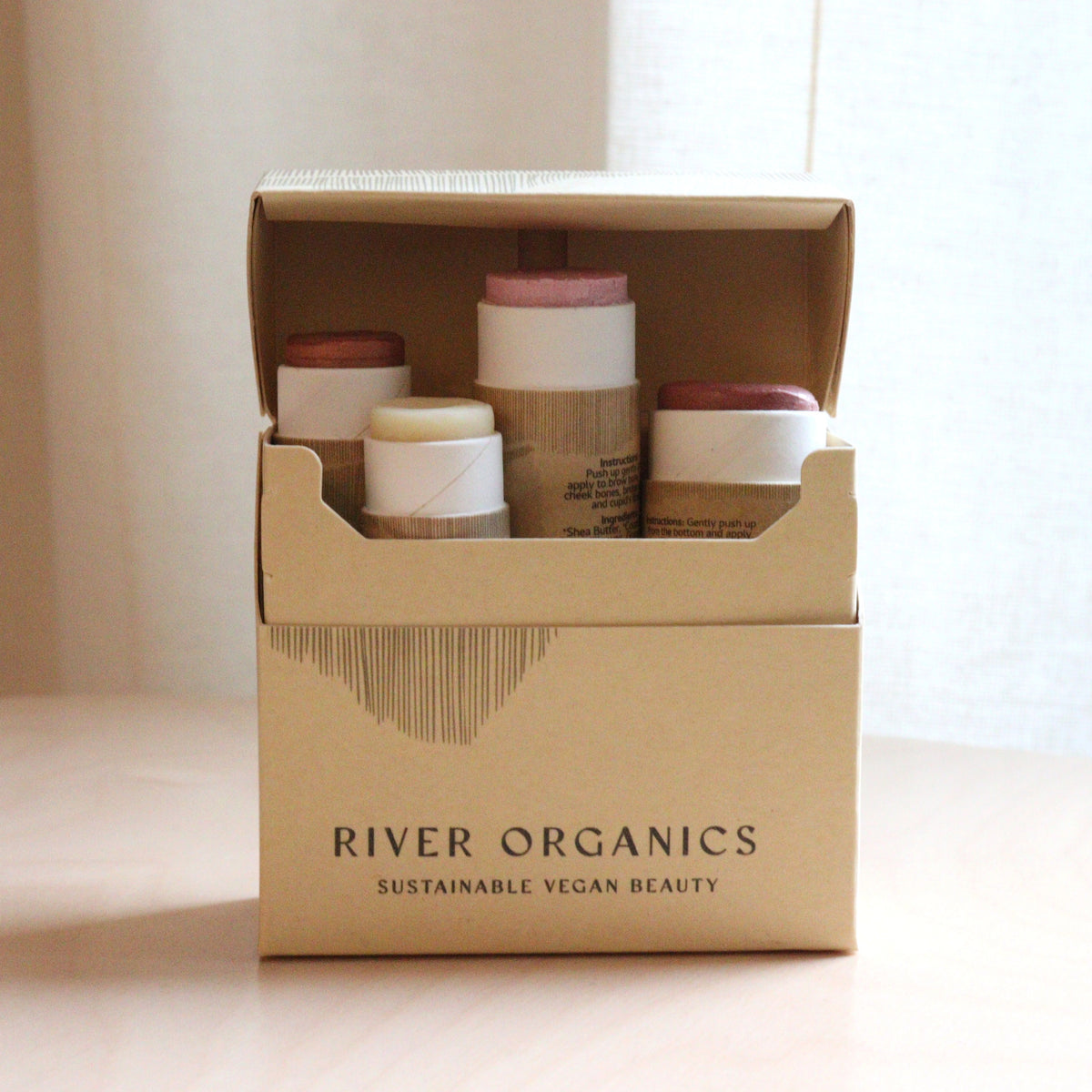 A cardboard box labeled "River Organics" sits on a table, open to reveal the Makeup Color Set by Tone with Blush, Highlighter & Two Lip Products—vegan beauty essentials in sustainable, natural shades.