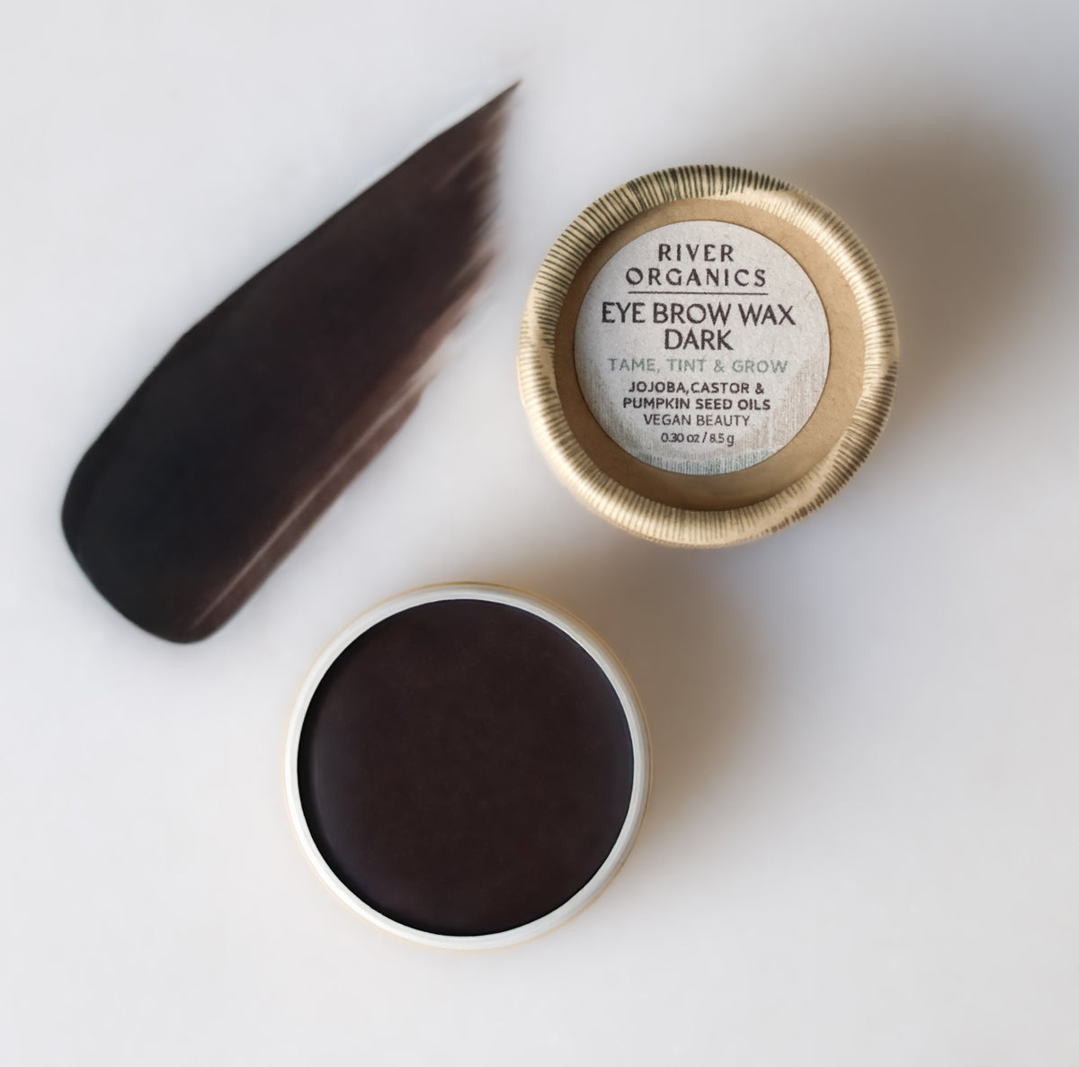 An open round container of River Organics Imperfect Brow Wax & Growth Serum reveals the dark brown vegan formula. The lid sits beside it displaying the label, and a swatch of the wax is smeared on a white surface.