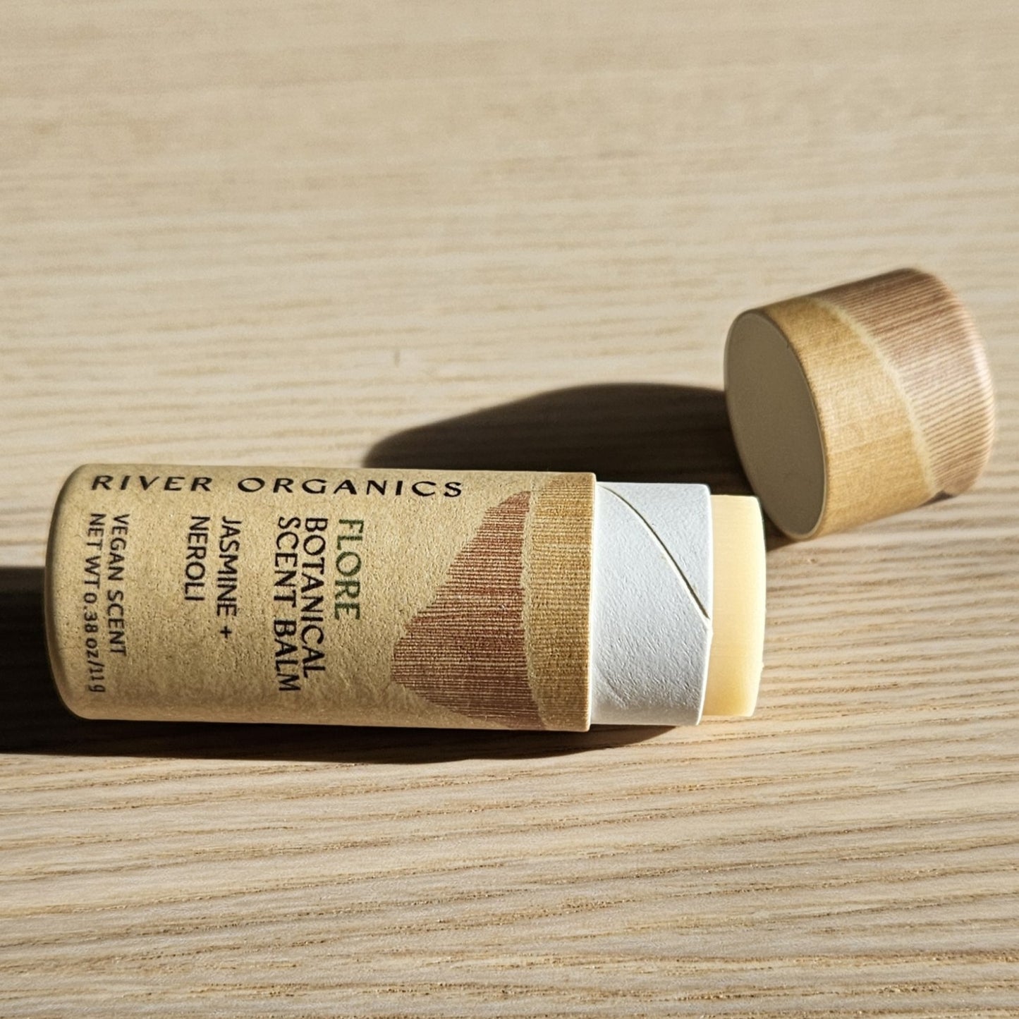 An open tube of River Organics Flore: Jasmine + Neroli Botanical Scent Balm sits on a light wood surface, its cap nearby. The brown, minimalist cardboard tube highlights this natural fragrance from River Organics.