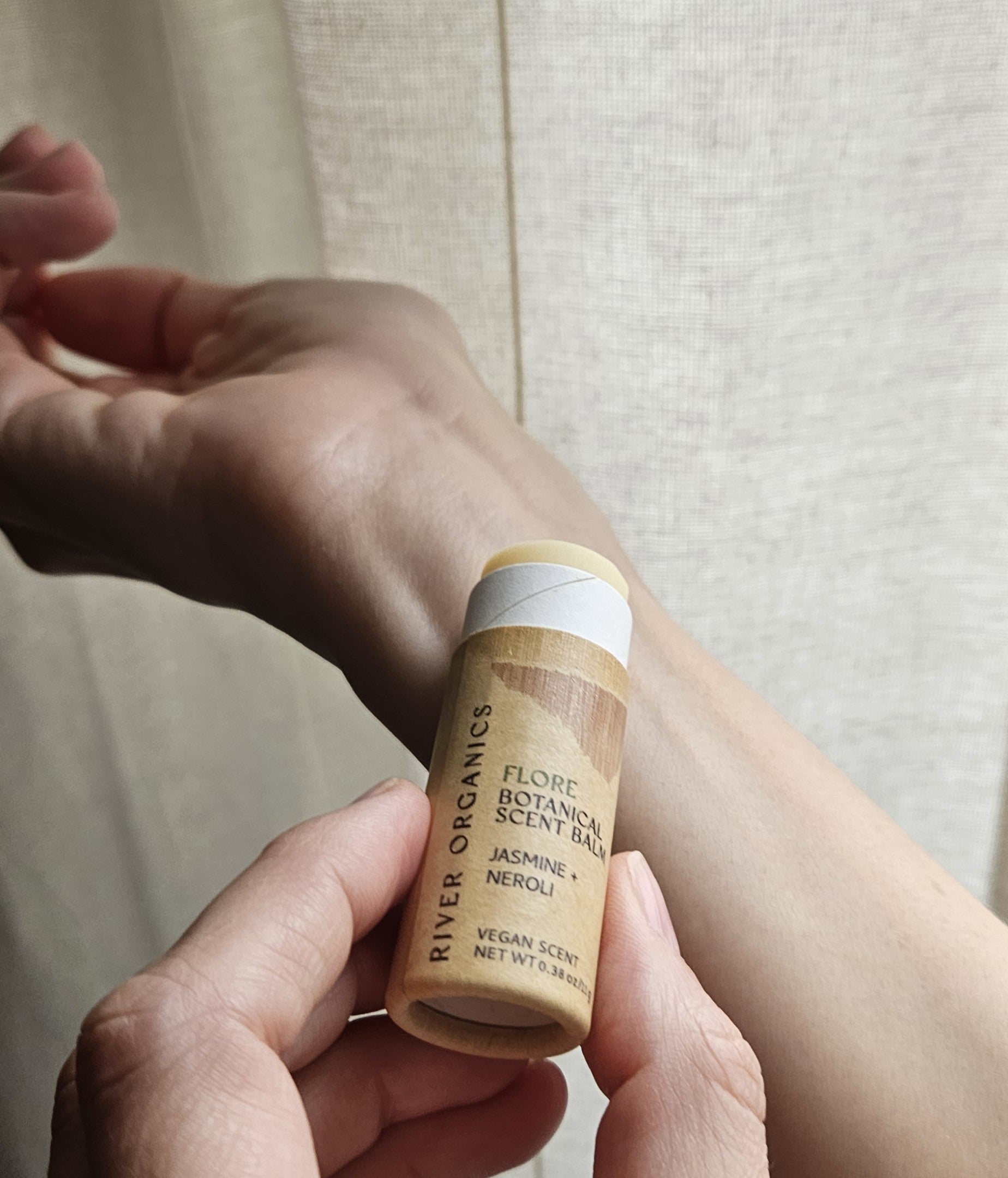 A hand holds River Organics’ Flore: Jasmine + Neroli Botanical Scent Balm near a wrist, ready to apply the natural fragrance. The label highlights jasmine and neroli; softly lit beige curtains are in the background.