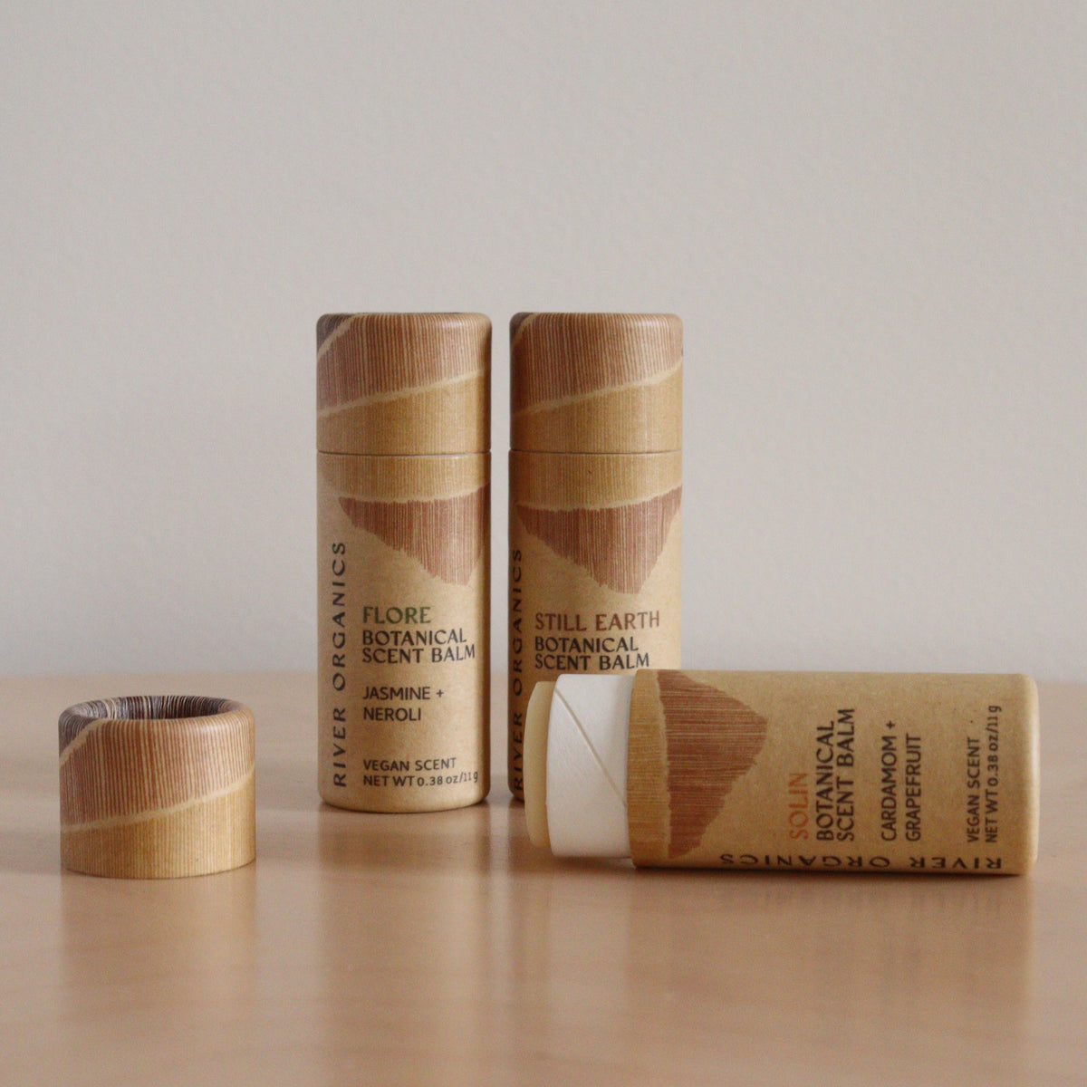 Three River Organics botanical scent balms, made with essential oils, are shown in cylindrical tubes with wood patterns—one open to reveal the white balm—all arranged on a light wood surface against a plain beige background.