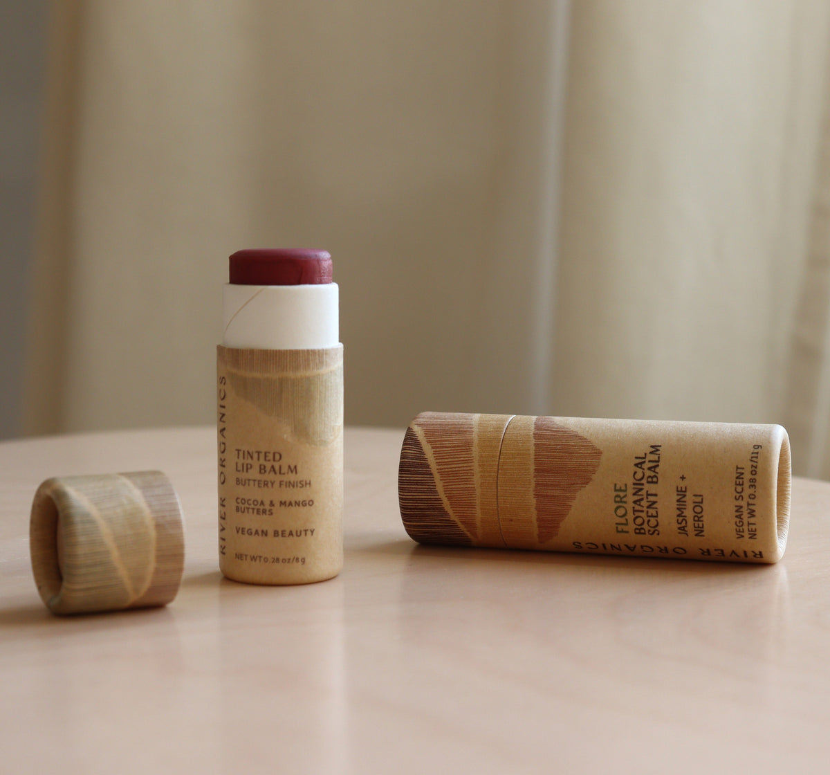 Two River Organics Lip + Scent Balm Bundle tubes rest on a table—one upright with its wooden cap nearby, another horizontal, showcasing nourishing organic oils and eco-friendly wooden packaging.